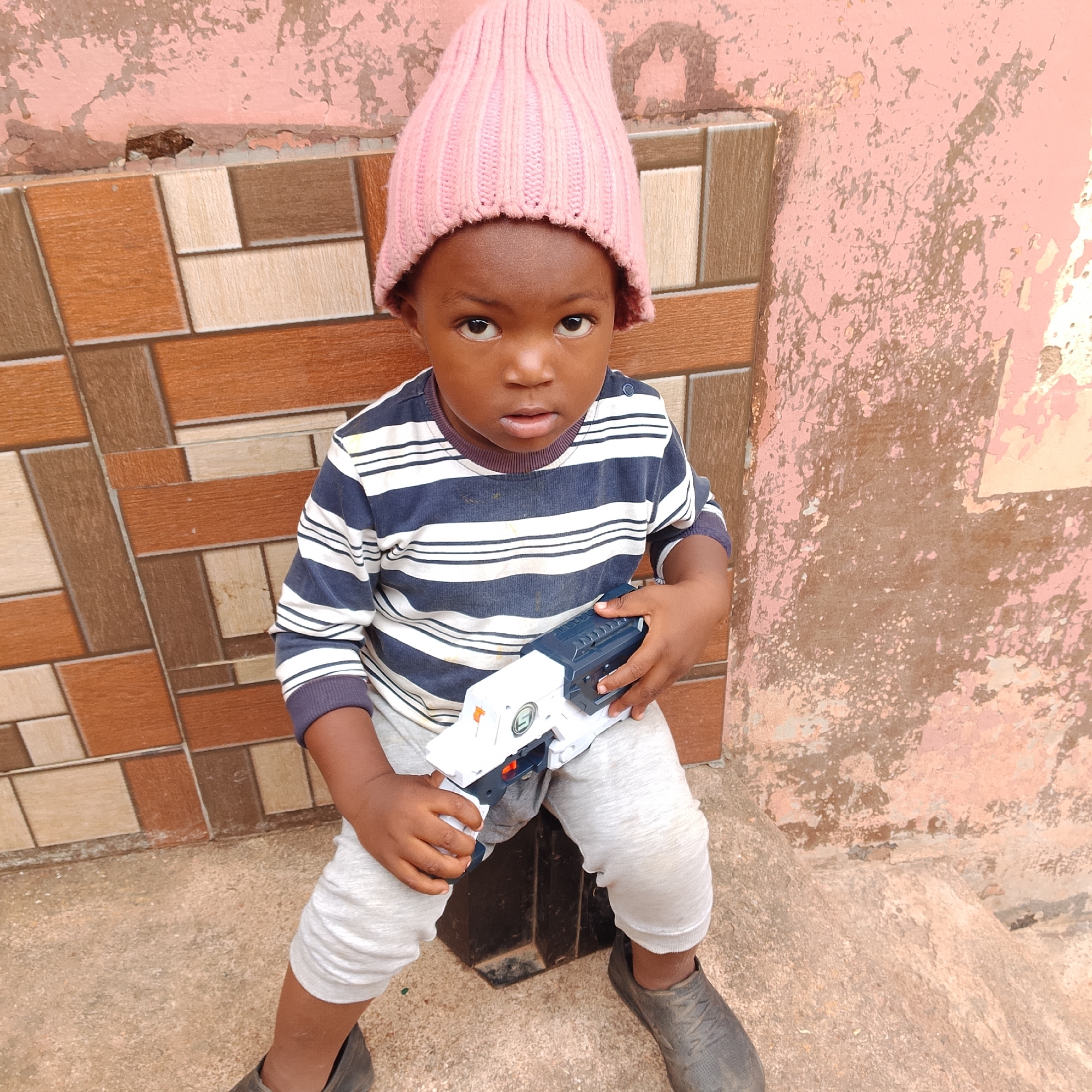 A young child, likely a toddler, is seated in front of a tiled wall and a textured, peeling pink wall. The child wears a pink knitted hat, a blue and white striped long-sleeved shirt, light gray sweatpants, and dark gray shoes. They are holding a white and dark blue toy gun in their hands, pointing it slightly forward. The child's expression is neutral, with eyes looking directly at the camera.
The background features a wall with alternating rectangular tiles in shades of brown and cream. To the right of the tiled wall is a section of pink wall with significant peeling and wear, revealing patches of darker material underneath. The ground is a rough, concrete-like surface. The scene appears to be outdoors, possibly in a residential area in Jos, Nigeria, given the architecture and weathered appearance of the walls. The lighting suggests it is daytime, with no strong shadows indicating a particular time of day. There is no visible text in the image.
