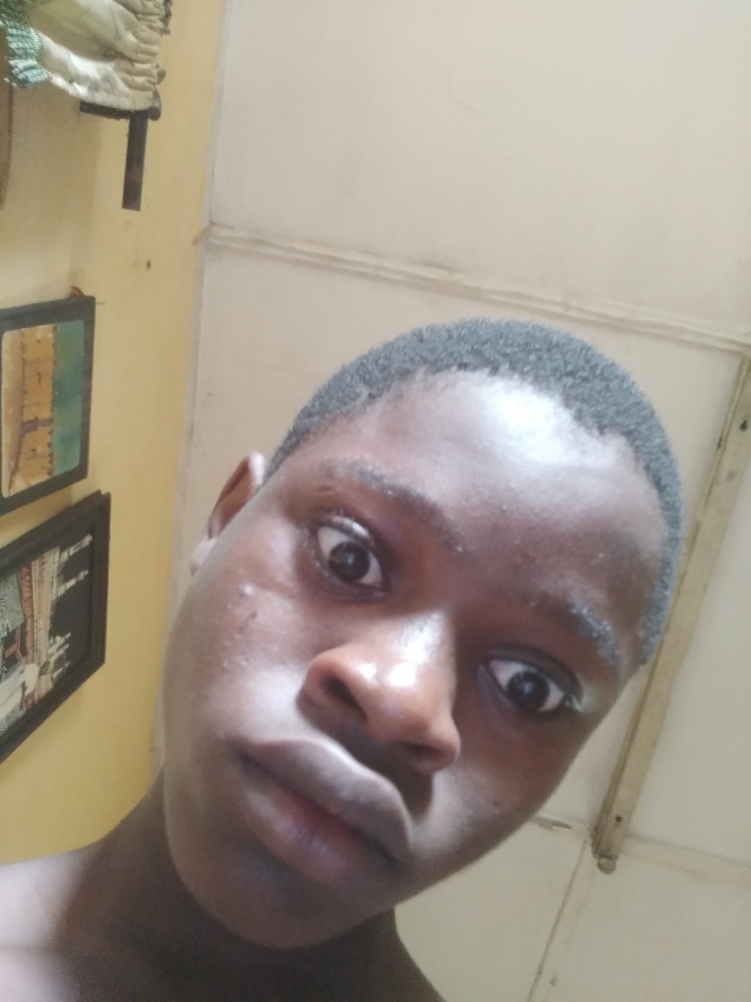 This close-up image features a young individual, likely male, with dark skin and short, dark, closely cropped hair. The person's face is angled slightly upwards and fills a large portion of the frame, looking directly into the camera with wide, expressive eyes and slightly pursed lips. Their skin appears somewhat shiny, possibly due to lighting or natural oils.
The setting is indoors, with a light yellow-painted wall visible behind the person on the left side of the frame. On this wall, two framed pictures are partially visible. The upper frame contains an image with shades of green and brown, resembling an abstract piece or a landscape. The lower frame, less distinct, shows what appears to be a black and white or sepia-toned scene, possibly featuring architectural elements or figures. Above these frames, in the top-left corner, a white, textured object, possibly a decorative fabric or ornament, is visible hanging on the wall, with a dark, narrow support extending downwards from it.
To the right and upper right of the person, a lighter-colored wall or ceiling, possibly off-white or cream, is present, featuring a seam or joint and a light-colored wooden or plastic vertical trim. The ambient lighting suggests the photo was taken during the daytime. The person is posing for a photograph, likely a selfie, with an expression that conveys attention or mild surprise due to their wide-open eyes. No specific activity is occurring beyond the portrait being taken. No visible text is legible in the image. The location context provided is Shangisha, Nigeria.