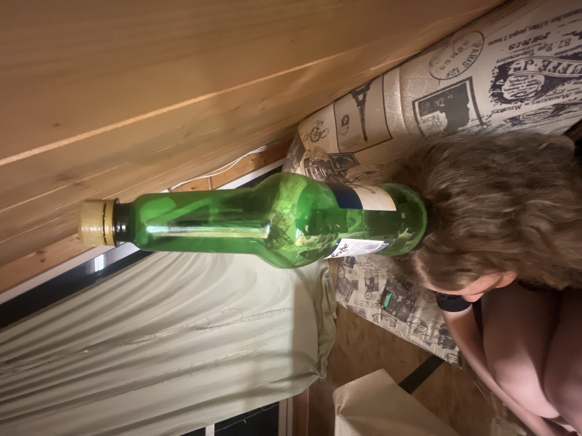 A photograph taken indoors shows a green glass bottle, laying horizontally, partially obscuring a person with curly brown hair. The person's face is not visible. They appear to be wearing black clothing and are seated. The setting seems to be an attic room, with wood paneling on the sloped ceiling. A window with a white curtain is visible on the left. The person is leaning against a padded headboard that has a Paris themed pattern. The bottle is positioned across the view, from the upper left to the center right. The information in the prompt about the city of Vasilyevo, Russia is not visible in the image.