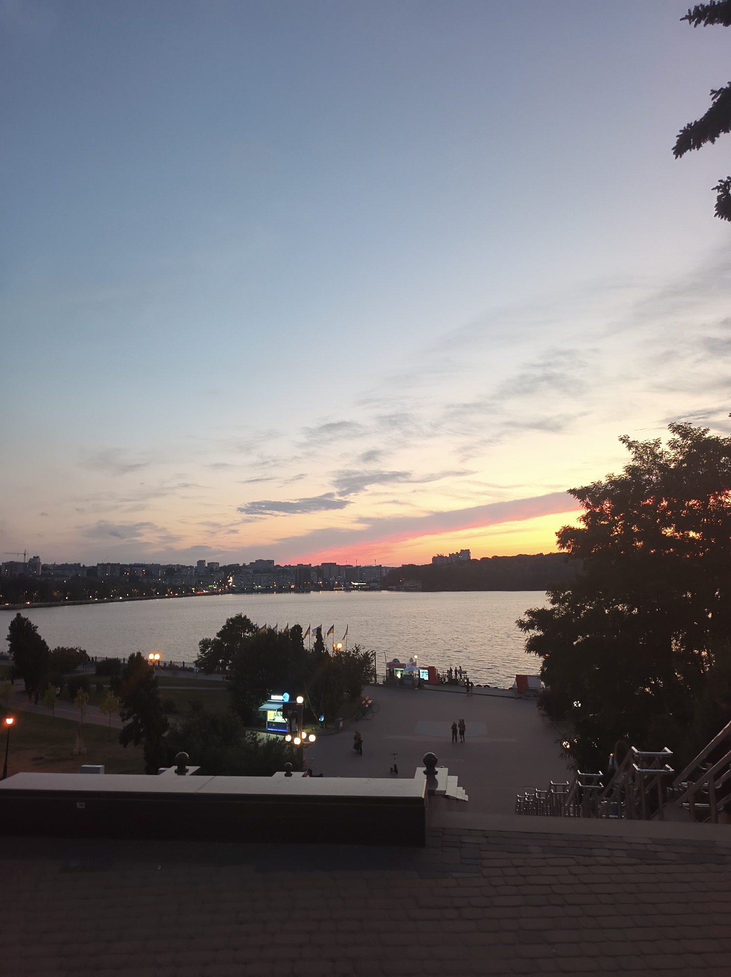 This is a wide-angle photograph taken at dusk, looking out over a body of water towards a cityscape. The sky is a gradient of soft blues and pale yellows, with streaks of pink and orange near the horizon indicating the setting sun. Silhouetted trees are visible in the foreground and middle ground, with one large tree dominating the right side of the frame.
The body of water appears to be a lake or river, with a distant shoreline lined with buildings and streetlights. Several flags are planted along the waterfront. The area immediately adjacent to the water features a paved walkway with some people strolling. There are also some kiosks or small businesses with illuminated signs.
In the foreground, there is a set of stairs leading down, with metal railings. The stairs are covered in dark, tiled paving. A low wall or bench is positioned to the left of the stairs. The overall impression is a public promenade or park area in a city.
The location is identified as Ternopil, Ukraine. This specific view is likely from a vantage point overlooking the Ternopil Pond (Teremno Lake), a large artificial lake in the center of the city, a popular recreational area. The cityscape in the background represents the urban area surrounding the pond.