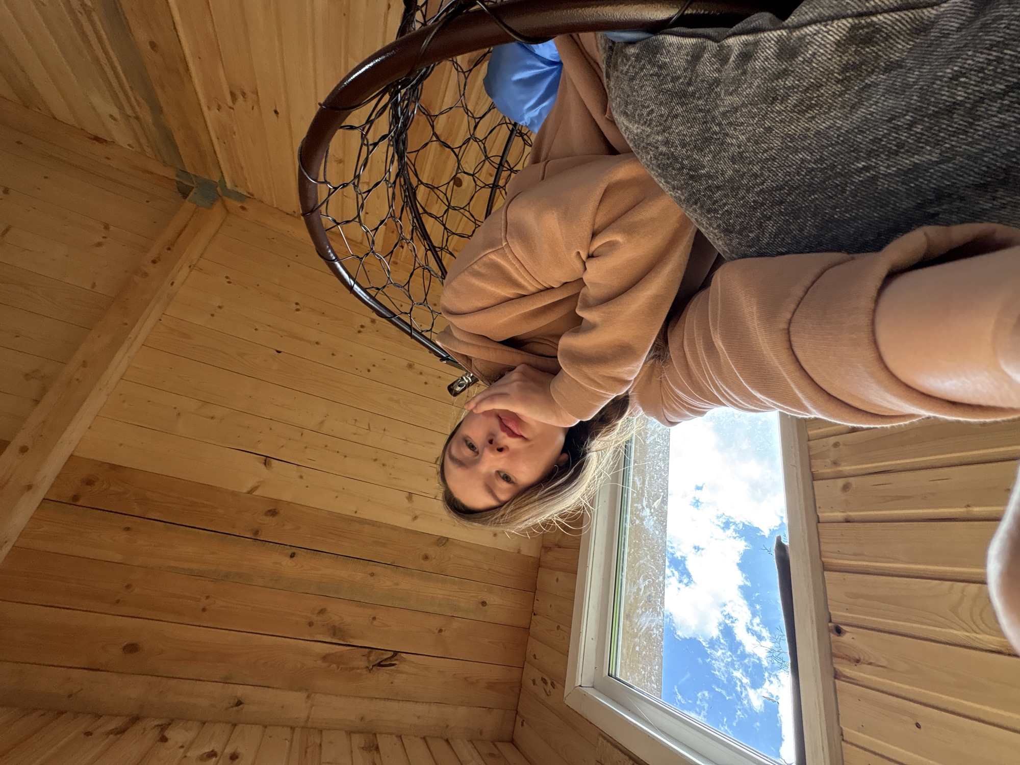 A woman with blonde hair sits in a hanging chair inside a wooden structure with a window. The chair is dark brown and has a mesh-like design. She is wearing a light brown hooded sweatshirt. A gray pillow is visible in the chair. Outside the window, the sky is blue with white clouds. The wooden walls and ceiling indicate the structure is possibly a cabin or similar dwelling. The location is Italmas, Russia.