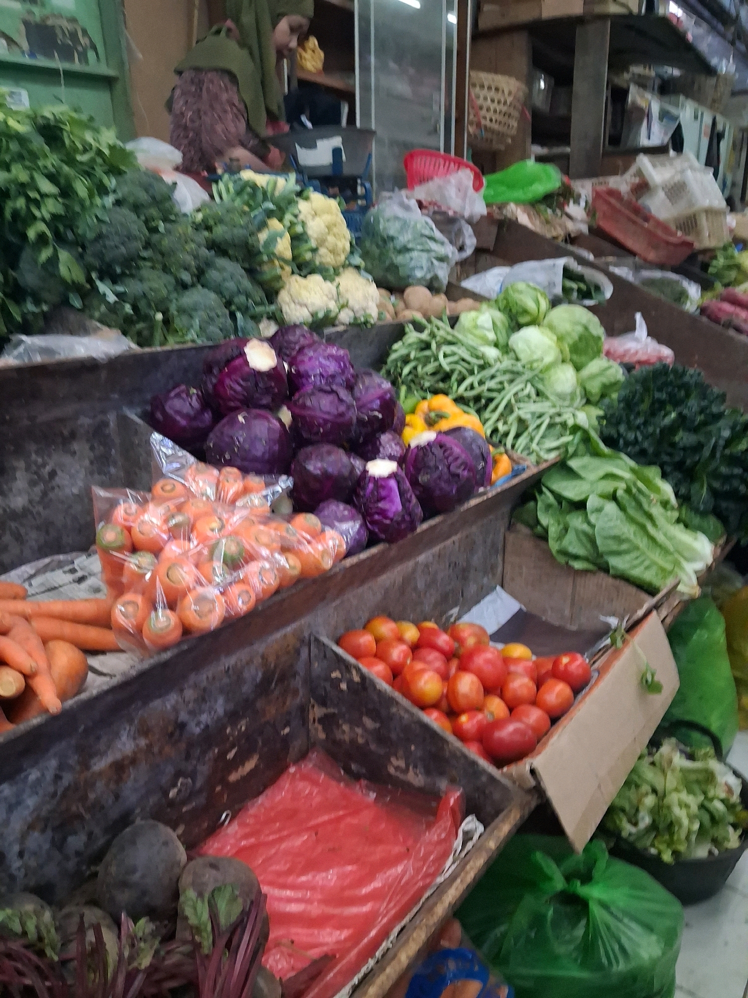 The image depicts a vibrant produce stall in a market, likely in the city of Surakarta, Indonesia, given the context. The stall is overflowing with a diverse array of fresh vegetables, arranged in wooden crates and on surfaces. In the foreground, a dark, rectangular crate contains several beets with their leafy tops, and a red plastic bag. To the right, another crate is filled with numerous red tomatoes and some yellow bell peppers, some of which are displayed in a cardboard box. Above this, a variety of green leafy vegetables, including lettuce and what appears to be long beans, are presented. Further back, a bin holds several heads of red cabbage, and a plastic bag contains a cluster of carrots. In the background, beyond the produce, a person with brown hair, wearing a green headscarf and a patterned top, is partially visible, seemingly interacting with the market environment. Other market stalls, baskets, and shelves are visible in the background, suggesting a bustling marketplace. The overall impression is one of abundance and freshness, typical of a local Indonesian market.