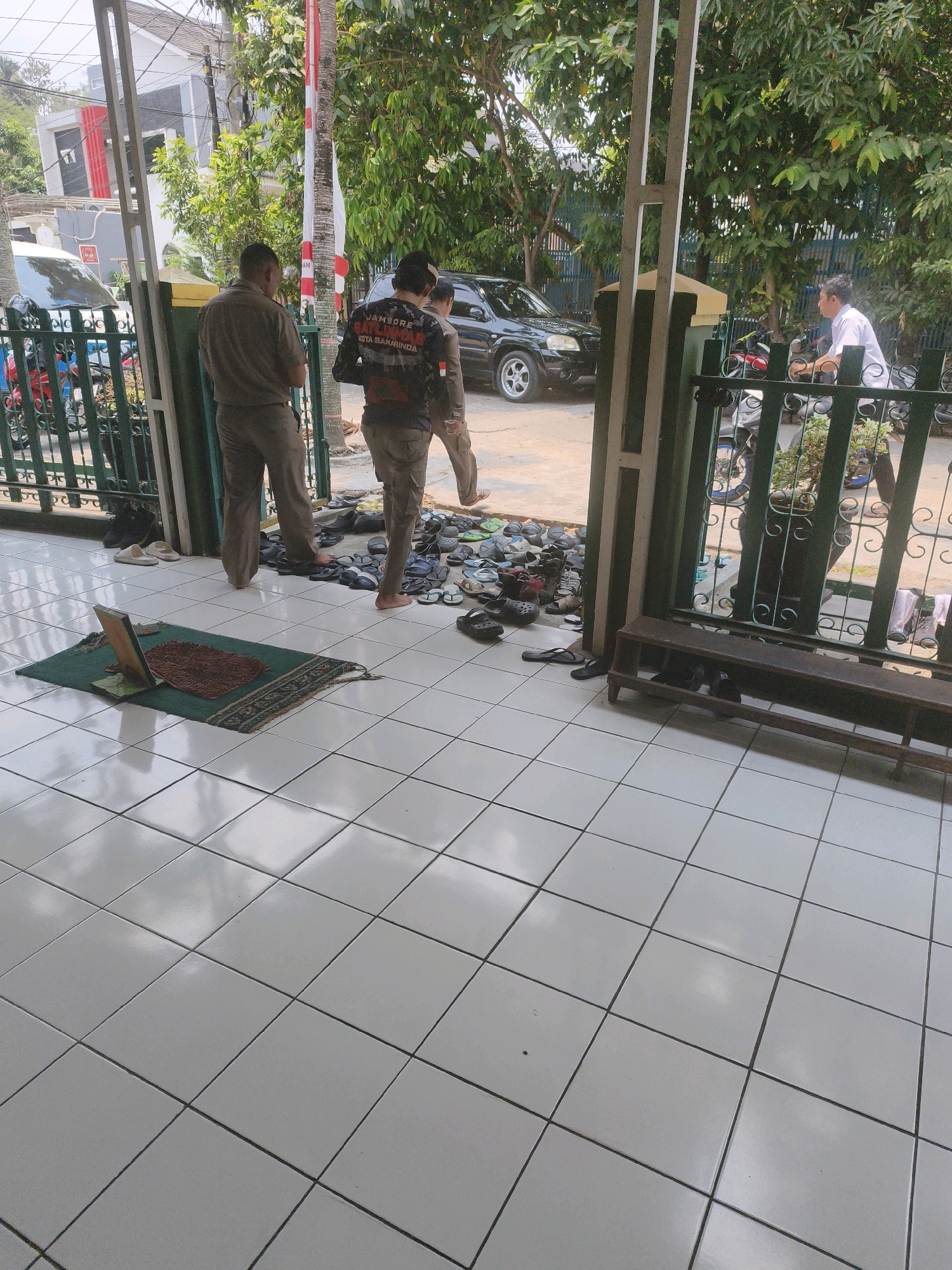 In Samarinda, Indonesia, a group of men are gathered near the entrance of a building, possibly a place of worship. One man in a gray uniform stands with his back to the camera, facing a collection of shoes and sandals arranged on the tiled floor. To his right, another man in a dark shirt with a graphic print and a backward-facing baseball cap is walking away, also towards the shoe pile. A third man, also in uniform, is walking in the same direction. In the background, a black SUV is parked. The tiled floor suggests an outdoor or semi-outdoor area. A prayer mat with a wooden stand is laid out on the left side of the frame. The scene appears to be in a residential or community area with trees and a white building visible in the distance.