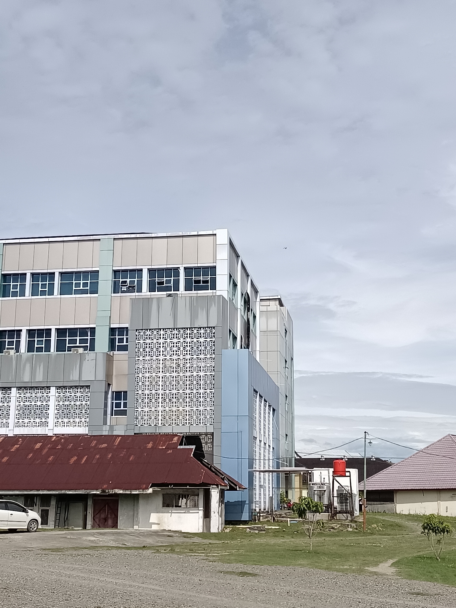 The image depicts a portion of a multi-story building with a light gray and beige facade, featuring numerous windows. One section of the building has a decorative patterned facade. In the foreground, there is a lower, older structure with a weathered, reddish-brown metal roof. To the right of this structure, a modern building with a light blue section is visible, along with other buildings and vegetation in the background. The sky is overcast. A white car is parked to the left of the older structure. A bird is visible flying in the distance.