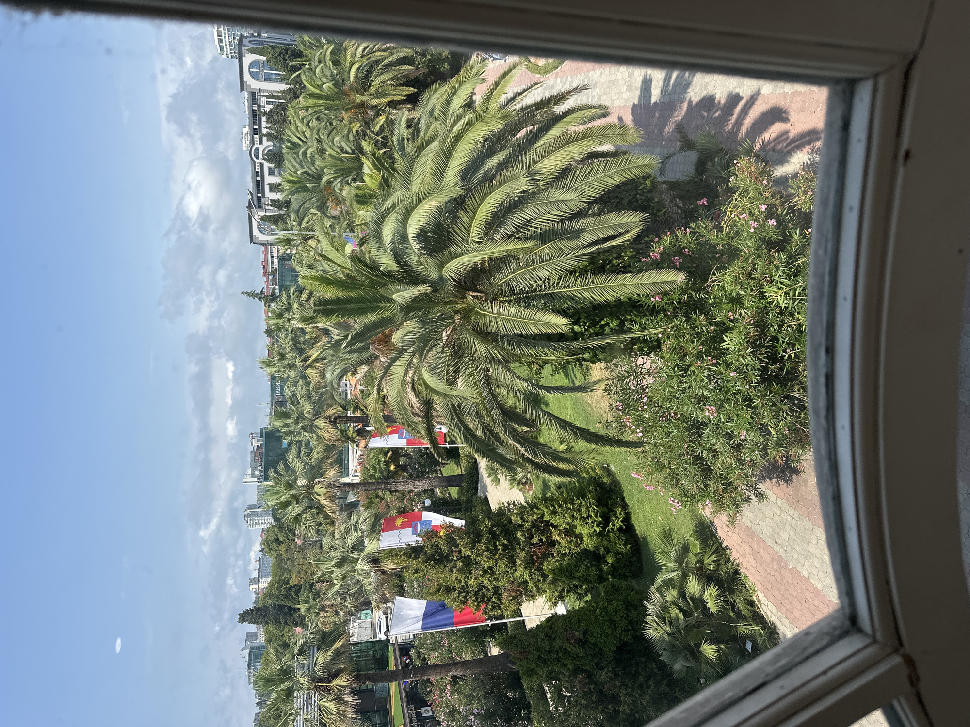 The image depicts a view of a resort or park area in Sochi, Russia, characterized by lush vegetation, palm trees, and a paved pathway. Several flags are visible, including the Russian flag and what appear to be regional or organizational flags, suggesting a formal event or establishment. Buildings, some with distinctive architectural features, are present in the background, indicating an urban setting within the resort landscape. The sky is partly cloudy, with a bright, sunny atmosphere. The foreground is framed by the edge of a window, suggesting the photo was taken from inside a building, looking out.