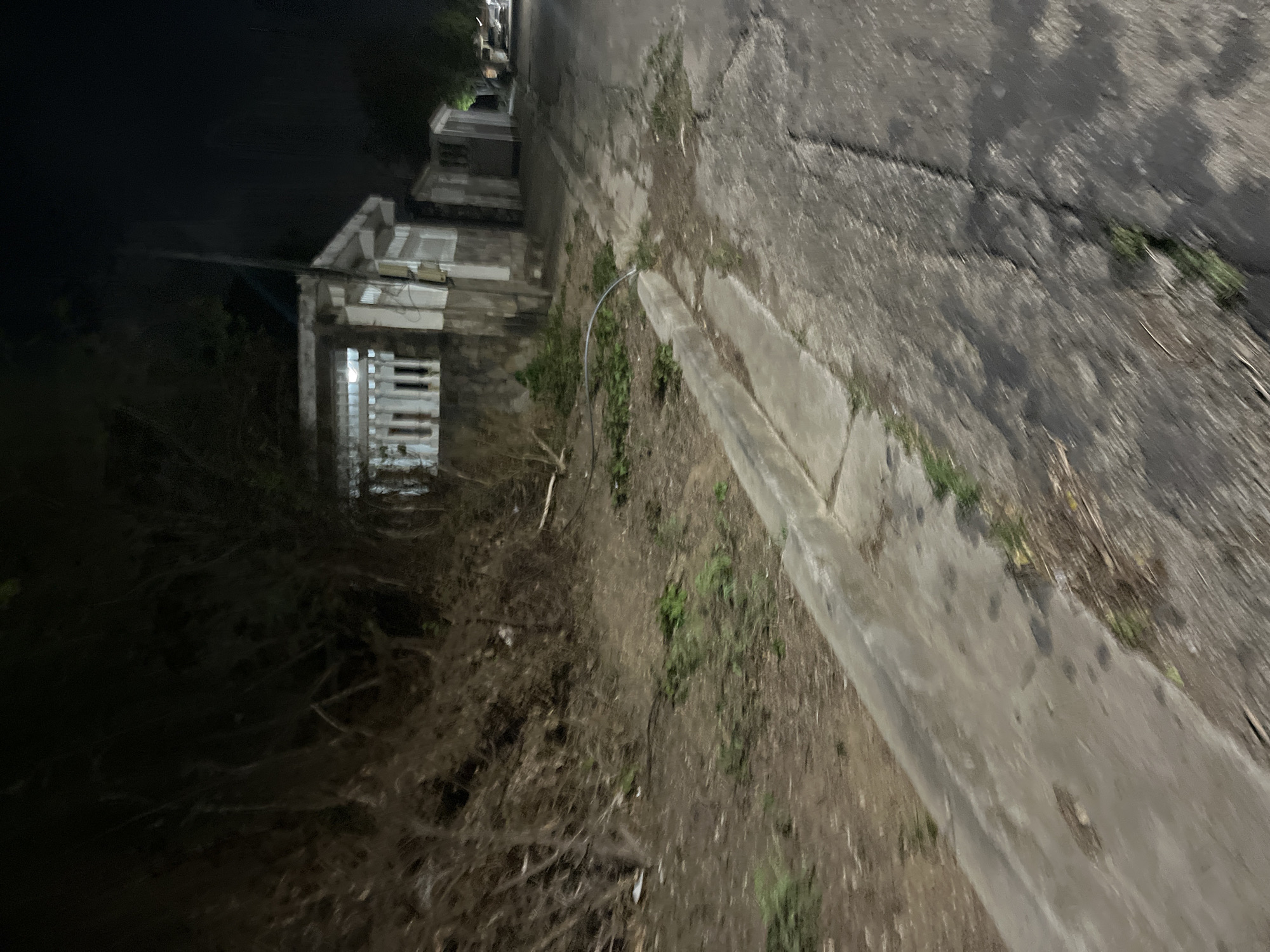The image depicts a dimly lit, outdoor scene at night, likely in a residential or semi-urban area. A concrete sidewalk with a curb runs along the left side of the frame, bordering a slope covered in dirt and debris, with overgrown vegetation and tangled branches. Beyond the slope, a small, light-colored building with decorative white vertical slats on its windows is visible. The building appears somewhat weathered. A flexible hose or pipe arcs from the ground near the curb towards the building. The ground adjacent to the sidewalk is a cracked asphalt road, also with scattered debris and patches of grass. Further down the road, another similar building structure is partially visible, and a vehicle's taillights are faintly discernible in the distance, suggesting it is moving away. The overall impression is one of a slightly neglected environment under artificial lighting. There are no people visible in the image.