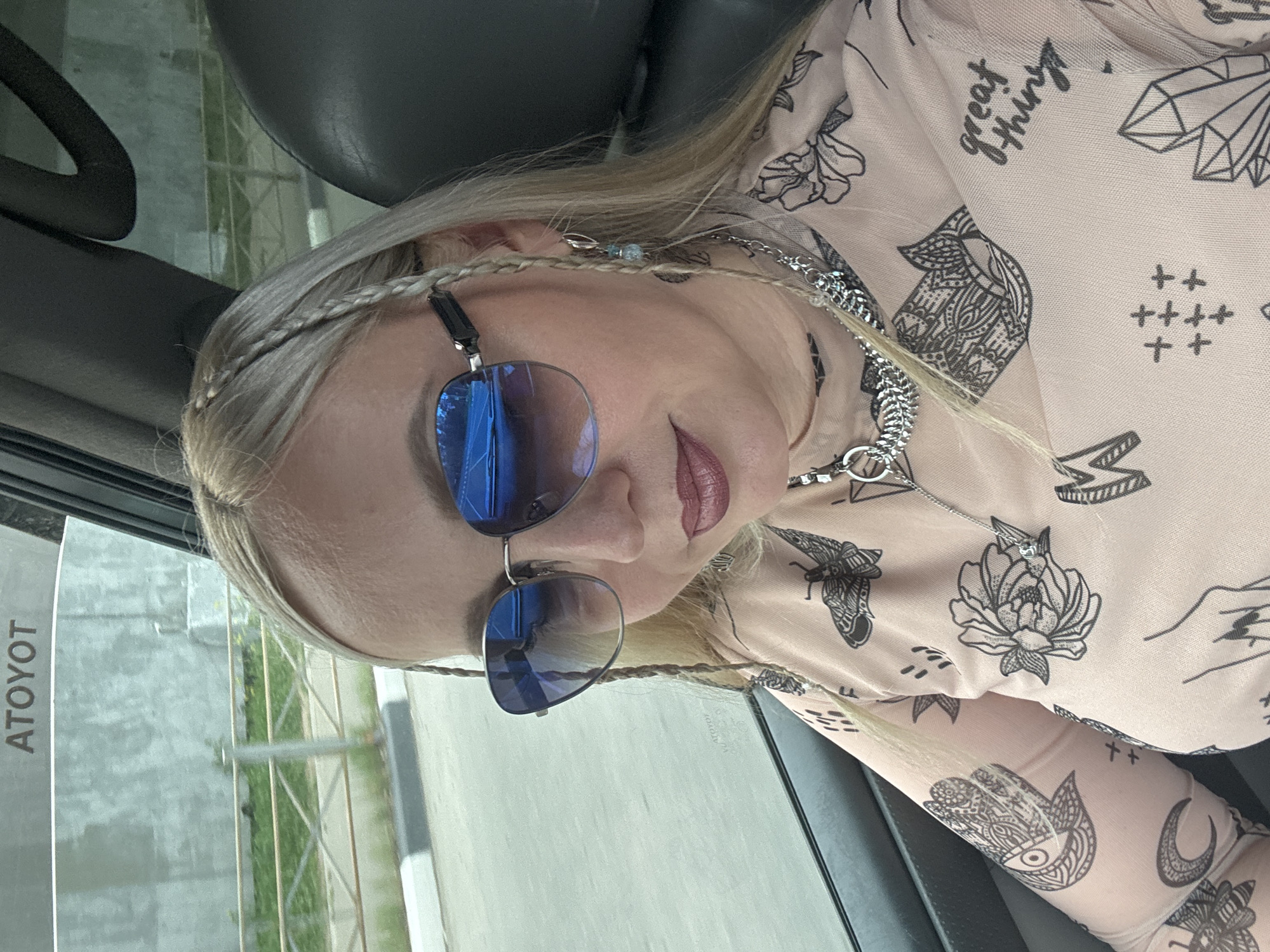 A blonde woman with braided hair and blue sunglasses is pictured inside a vehicle. She is wearing a light pink, long-sleeved shirt with black line drawings of butterflies, flowers, hands, and crystals. She has on silver necklaces and earrings. The vehicle appears to be a car or van. Through the window, a grey wall, a metal fence, and greenery can be seen, suggesting an outdoor environment. The text "ATOYOT" is visible on the window, potentially indicating a brand or location. The scene is shot from a slightly elevated angle, capturing the woman's upper body and face. The background details are blurred, focusing attention on the subject.