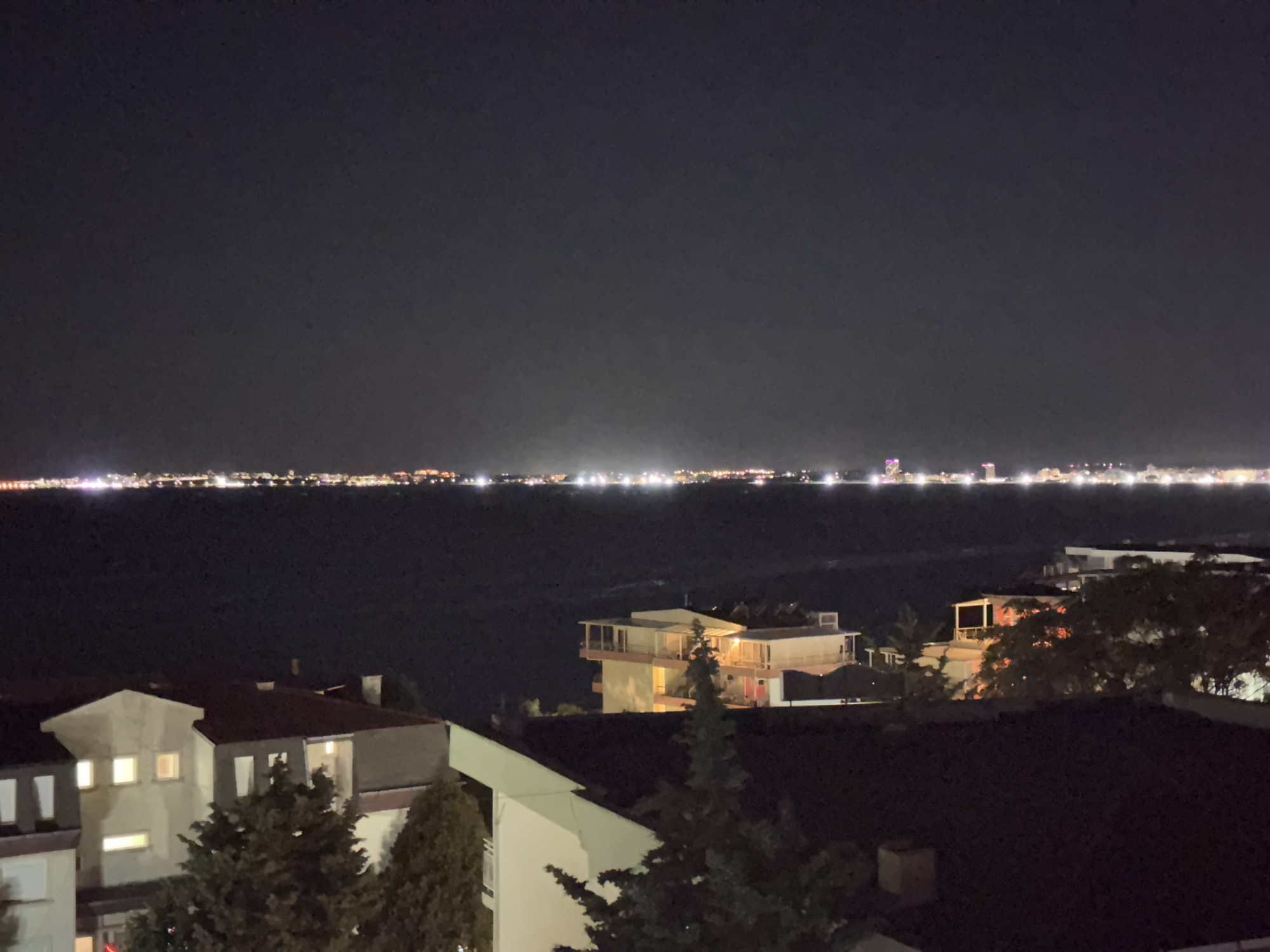 The image shows a night view overlooking a body of water with a distant city skyline illuminated by lights. In the foreground, there are several buildings, including houses and a multi-story structure with balconies. Trees are also visible, some of which appear to be evergreen. The water is dark, reflecting some of the distant lights. The sky is dark and clear, with no visible celestial bodies. The overall scene suggests a coastal city at night, possibly Sveti Vlas, Bulgaria, given the provided location context.