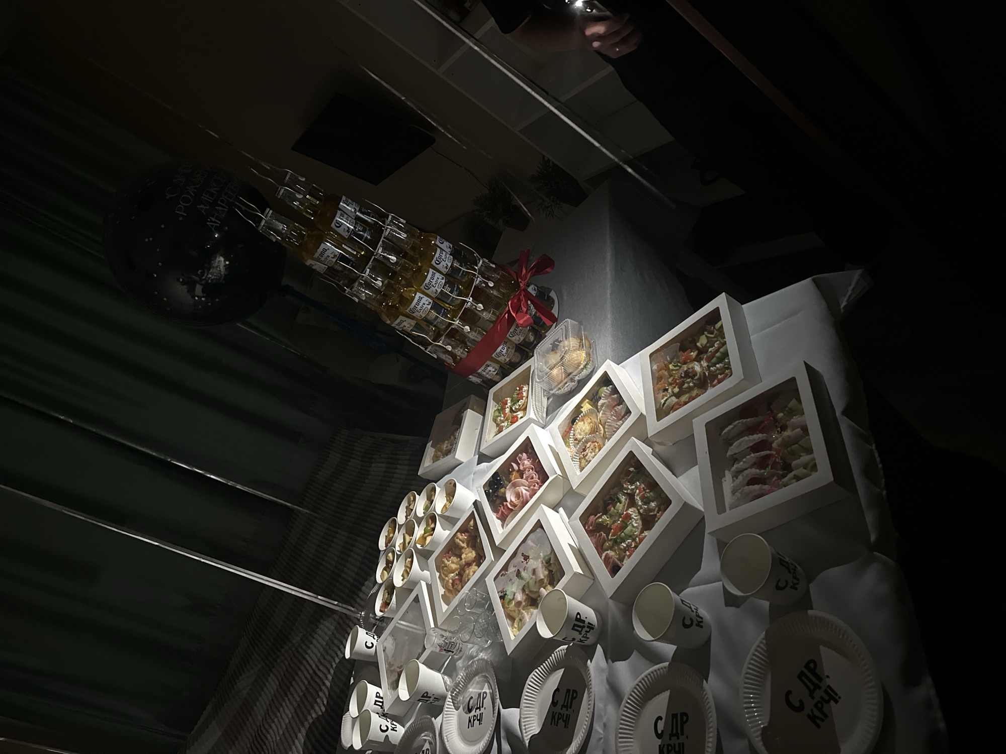The image depicts a dimly lit indoor scene, possibly a party or gathering, set against a backdrop of dark green curtains and a striped bedspread or couch. The primary focus is a table laden with food and drinks, arranged for a celebration.
In the foreground, there are several white paper plates with black text that appears to be in Cyrillic. Adjacent to the plates are white paper cups, also with similar Cyrillic lettering. Scattered amongst the plates and cups are various food items presented in white cardboard boxes with clear plastic lids. These boxes contain an assortment of appetizers, including what appear to be mini pizzas or bruschettas with toppings like vegetables and possibly cheese, as well as other small, colorful bites that could be canapés or hors d'oeuvres. Some boxes contain pasta or noodle salads.
A prominent feature of the scene is a tower constructed from bottles of Corona Extra beer, which is wrapped with red ribbon and fairy lights. This beer tower is positioned towards the right side of the table. Above the beer tower, a black balloon with white polka dots and Cyrillic lettering is visible. The lettering on the balloon seems to read "С Днем Рождения" (Happy Birthday) and a name or inscription.
Further back on the table, more cups and small containers of food, possibly fruit or dessert portions, are visible. Two small, clear shot glasses are also present. The overall impression is a catered or prepared spread for a festive occasion.
The lighting is dramatic, with strong shadows cast across the table and the background, suggesting the use of artificial light. The environment appears to be a private residence or a small event space.
Regarding the location of Perm, Russia, there are no direct visual cues in the image to confirm this specific city. However, the Cyrillic script on the plates, cups, and balloon strongly indicates a Russian-speaking locale. Without additional context, pinpointing the exact city of Perm based solely on this image is not possible. The scene itself is a generic representation of a party setup.