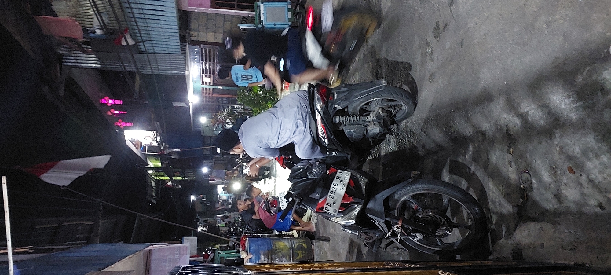 The image depicts a nighttime street scene in Jayapura, Indonesia, characterized by dimly lit conditions and a busy, residential atmosphere. In the foreground, two parked motorcycles are visible. The one on the left is black and red with a license plate reading "PA 2926 RJ". The rider on the motorcycle to the right is wearing a light blue shirt and a black cap, facing away from the camera. Further back, other individuals and motorcycles are present, with some people appearing to be socializing or moving around. The environment includes buildings with corrugated metal roofs and balconies adorned with flags and what appear to be decorative lights. The ground is a paved surface, with some debris visible. The overall impression is of a casual, everyday gathering in a neighborhood setting at night.