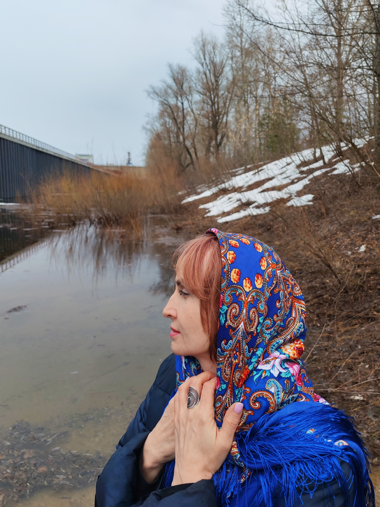 A woman stands near a body of water in what appears to be a natural setting within an urban environment. The location is Chaikovsky, Russia. The woman is wearing a blue patterned headscarf (Pavlovo Posad shawl) with fringe. She has reddish-blonde hair and a ring on her finger. She is wearing a dark blue jacket and has her hands near her neck. To the left is a grey bridge structure over water. The water surface has some reflections of the sky and surrounding structures. The ground on the right is covered in vegetation and some patches of snow. Bare trees are in the background. The sky is overcast.