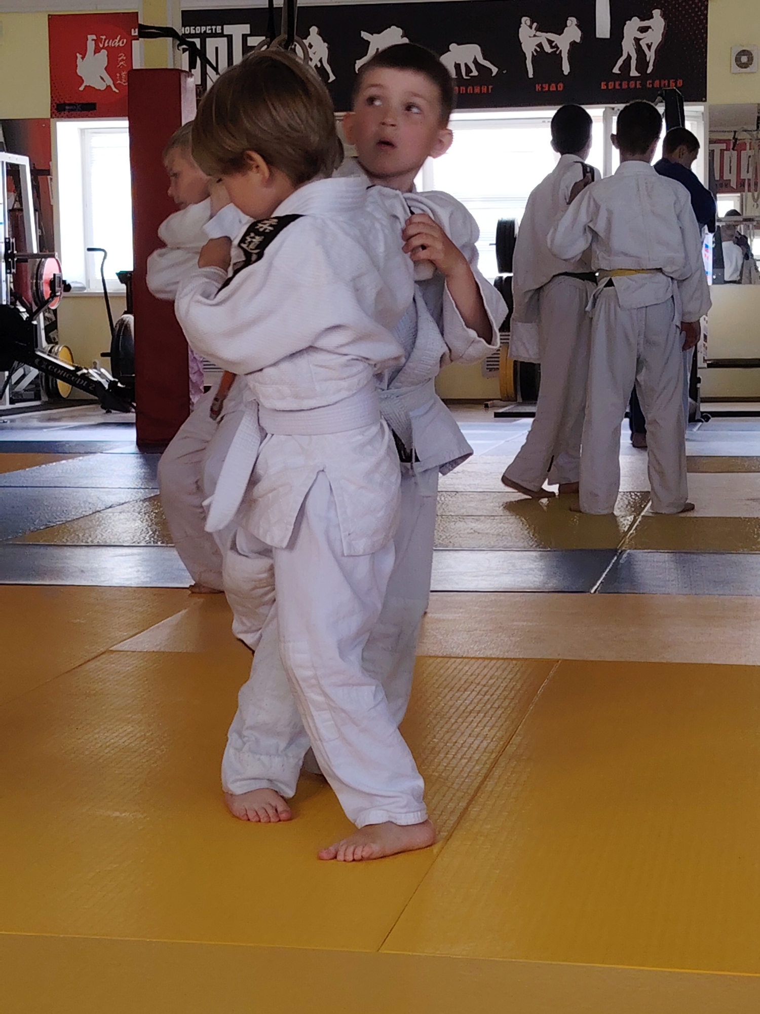 The image captures a martial arts training session involving several young children, likely in a dojo or gym located in Chaikovsky, Russia. In the foreground, two children dressed in traditional white judo gis are practicing a grappling technique. The child closest to the viewer has their back partially turned, revealing a white belt and bare feet on the yellow training mat. The other child in the foreground looks slightly upwards, mouth agape, possibly reacting or observing.
The background shows more children in white gis, some facing away, indicating a larger class. One child in the mid-ground wears a yellow belt. The floor is covered with a combination of yellow and blue-grey interlocking mats.
The room is illuminated by natural light entering through a window on the left. Various gym equipment, including one marked "CONCEPT 2," is visible near the window. The walls are adorned with martial arts-themed posters. A prominent red poster on the left features "Judo" in both English and Japanese script, alongside a silhouette of two figures. A long black banner above the background participants displays various martial arts silhouettes and text in Russian, including "БОРЬБА" (Struggle/Wrestling), "КУДО" (Kudo), and "БОЕВОЕ САМБО" (Combat Sambo), suggesting a diverse combat sports facility. A large mirror reflects parts of the training area and more children in the background. The overall scene conveys an active and dedicated training environment.
