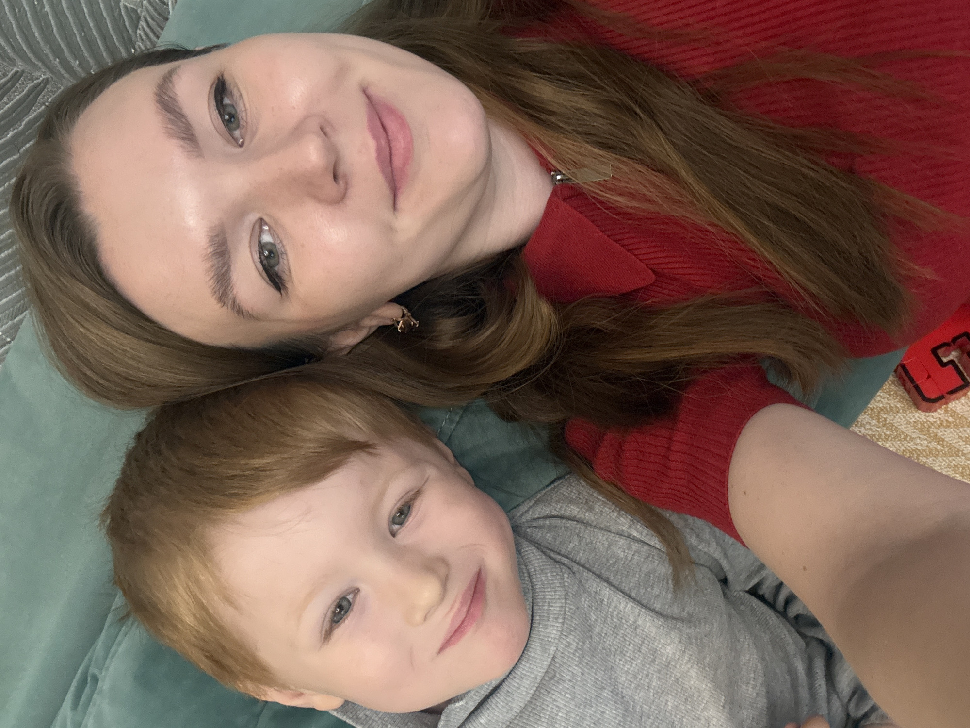 A woman and a young child lie together on a light blue surface, likely a bed or sofa. The woman, positioned at the top of the frame, has light skin, brown hair, and is wearing a red sweater. She is smiling and looking directly at the camera. The child, located below her, has fair skin, red hair, and is wearing a gray sweatshirt. The child is also smiling at the camera. The background includes a textured light blue surface and, in the top left corner, hints of a striped or patterned wall. A red and black object is visible to the right of the subjects. The image is a close-up shot. No information indicates the location is Izhevsk, Russia.