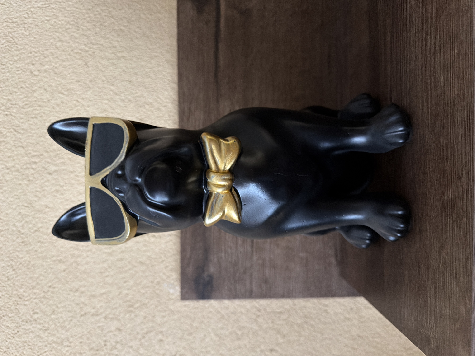 A black, glossy dog figurine, possibly a French Bulldog, is lying down. The figurine has a gold bow tie and gold-rimmed sunglasses. It is on a brown shelf, possibly made of wood, and next to a beige or light tan wall. The location is not discernible from the image.