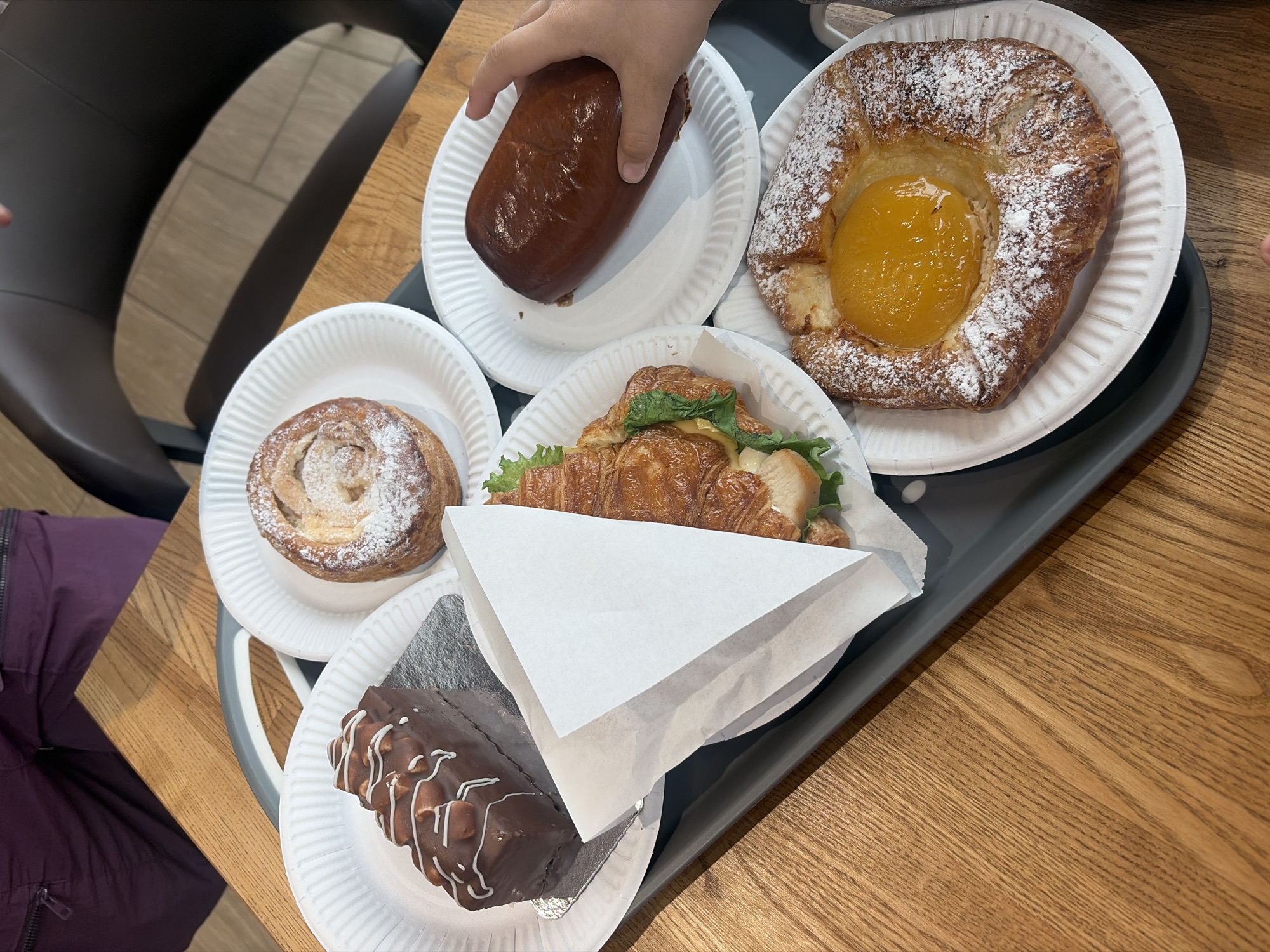 The image shows a grey tray on a wooden table holding five white paper plates with baked goods. Clockwise from the top left, the plates contain: a shiny, oblong pastry being held by a child's hand; a round pastry with a yellow filling, dusted with powdered sugar; a croissant sandwich partially obscured by a white paper wrapper; and a chocolate cake slice with white icing drizzled on top. A round pastry dusted with powdered sugar sits on a plate between the chocolate cake and the oblong pastry. A person wearing a purple jacket is partially visible on the left side of the image. A black chair is visible in the upper left corner. The scene appears to be indoors, possibly a bakery or cafe. The location is presumed to be St. Petersburg, Russia.