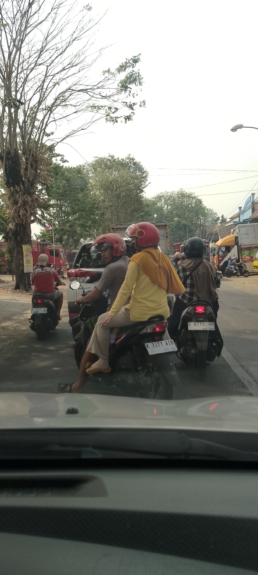 In the city of Panjang, Indonesia, on a cloudy day with a bright sky, a traffic jam is captured on a paved road. A group of people are on motorcycles, many wearing helmets. In the foreground, a motorcycle with a license plate reading "K 2477 AIB" is visible. The rider is wearing a gray shirt and a red helmet, while the passenger is wearing a yellow shirt and a light brown hijab, with tan pants. Another motorcycle behind them has a license plate "K 6774 RR". On the left side of the image, a motorcycle with a red shirt rider and a white helmet is seen from behind. The background features a variety of trees, buildings, and other vehicles including trucks. A tall, bare tree is prominently displayed on the left side of the image. A street lamp is visible in the upper right corner.