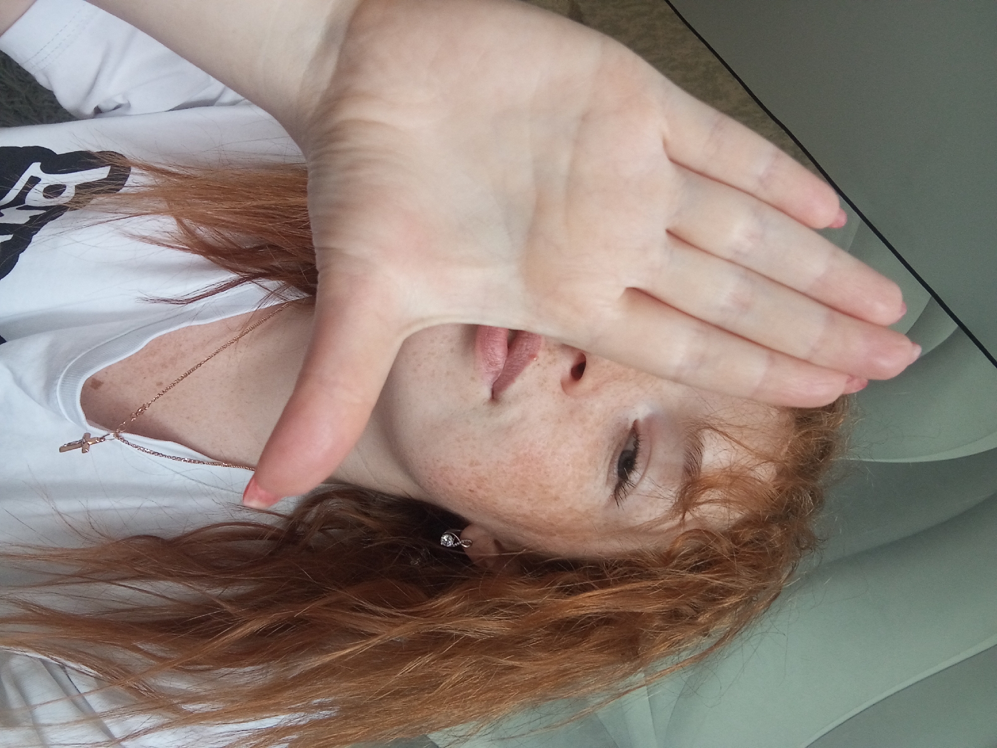 A fair-skinned person with red hair, numerous freckles, and a gold cross necklace is lying down and covering part of their face with their hand. They are wearing a white shirt with a black graphic. A silver earring is visible in their left ear. They appear to be indoors, possibly in a car. The specific location in the city of Unknown, Unknown is not discernible from the image.