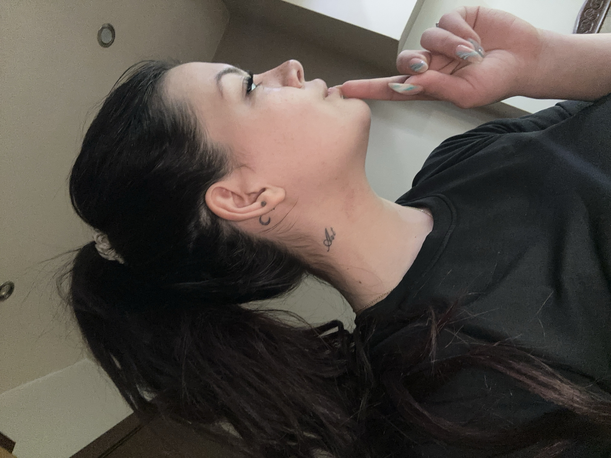 A woman with dark hair in a low ponytail, wearing a black top, is positioned in the lower right of the frame. Her head is tilted back and to the left, with a finger touching her lips. She has tattoos on her neck and behind her ear. The background is a plain wall with a recessed ceiling light. The location is uncertain and cannot be determined to be Sliven, Bulgaria, based solely on the visual information provided.
