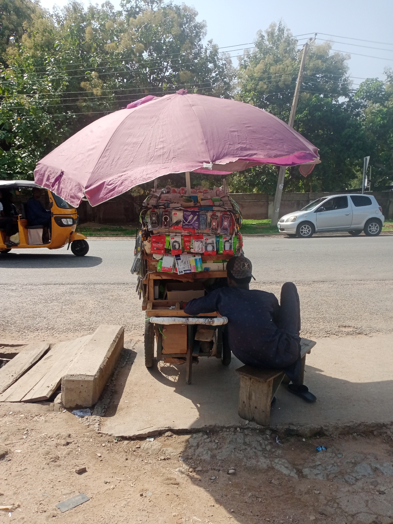 A man sits on a wooden stool on a paved sidewalk next to a street. He is wearing a dark blue outfit and a checkered cap. He is facing away from the camera, looking towards a street vendor's cart. The cart is covered by a large, pink umbrella and is laden with various mobile phone accessories, such as chargers, earphones, and phone cases. These items are neatly displayed in their packaging.
Behind the cart, a yellow auto-rickshaw is driving on the street. Further down the street, a silver hatchback car is parked. The scene appears to be in a commercial area, likely a roadside market or street vending spot in Jalingo, Nigeria. The weather is bright and sunny, suggesting it is daytime. The man on the stool seems to be taking a break or waiting for customers. The overall atmosphere is typical of a bustling street scene in a Nigerian city. There is no visible text in the image.