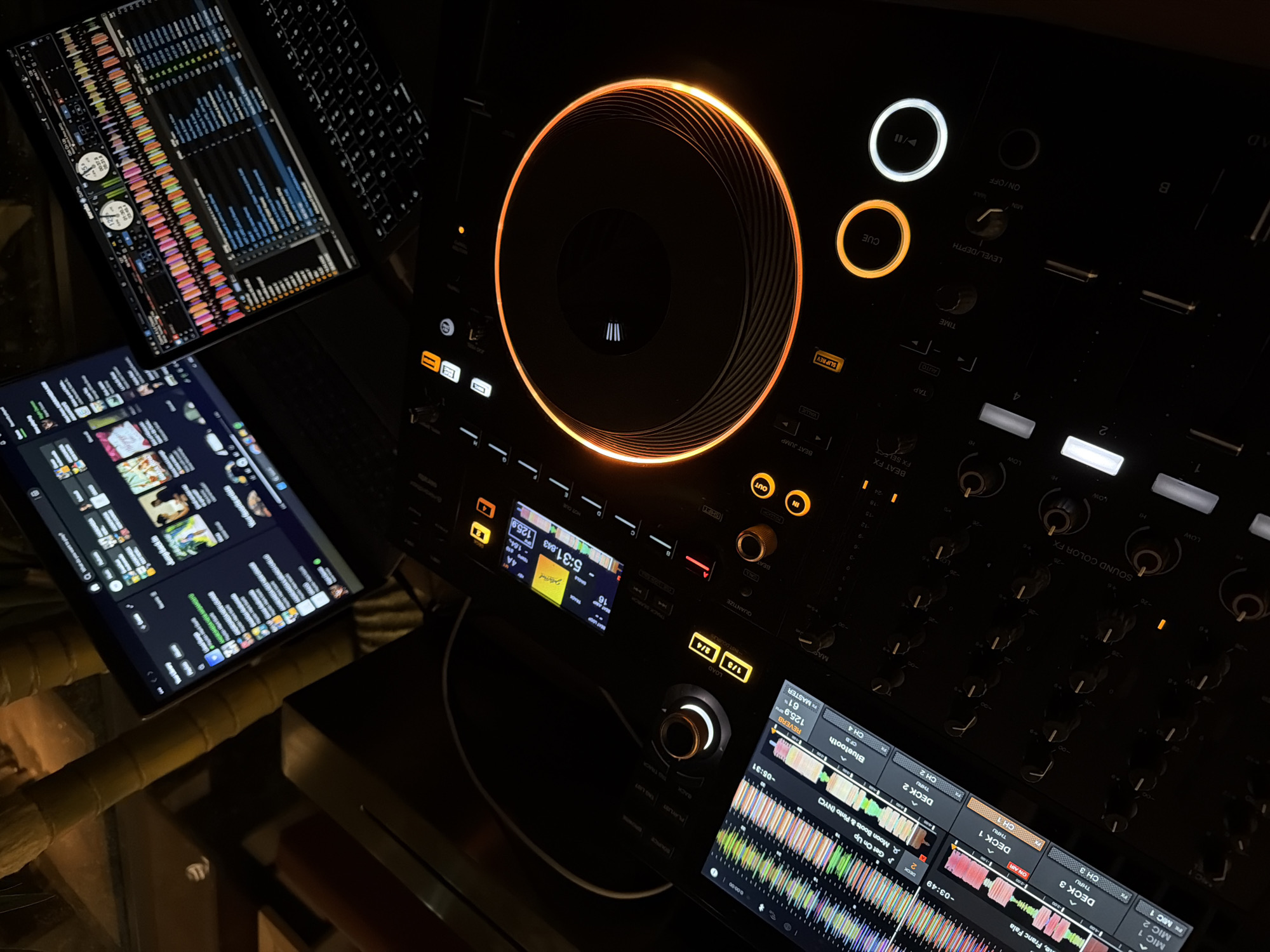 The image displays a professional DJ setup bathed in the warm glow of its operational lights, indicating a dark indoor environment, likely at night, in Budva, Montenegro. The central piece of equipment is a DJ controller, prominently featuring a large, circular jog wheel illuminated by a striking orange light. Surrounding it are various buttons and knobs, emitting white and orange lights that highlight functionalities such as "CUE," "IN," "OUT," "SHIFT," "SLIP REV," "VALUE," "ON/OFF," "LEVEL/DEPTH," "TIME," and "BEAT FX."
To the left, a laptop or tablet screen presents intricate music production software, showcasing vibrant, multi-colored audio waveforms, vertical level meters, and detailed track information. A keyboard is partially visible next to it. On the right side of the main controller, another screen displays waveforms for "DECK 1" and "DECK 2," alongside track titles such as "Moon Boots & Pinto (NYC) - Get On Up" and "Me, Franc Fala." Time displays like "-03:49" and "-05:31" indicate playback progress.
Further visible text on the equipment includes numerical BPM readings like "125.9" and "126," "61%" (possibly gain or volume), and control labels such as "QUANTIZE," "LOAD," "REVERB," "MASTER," and "SOUND COLOR FX." Channel labels ("CH 1" through "CH 4") and deck assignments ("DECK 1" through "DECK 3") are also visible. The screens mention "Bluetooth" connectivity and UI elements like "PLAYLIST," "ENG LIST," and "BACK." The phrase "serato DuPo" is partially visible in the upper-left screen, likely referring to Serato DJ Pro software. The scene captures an active DJ session or the preparation for one, characterized by the intricate interplay of light and technology.