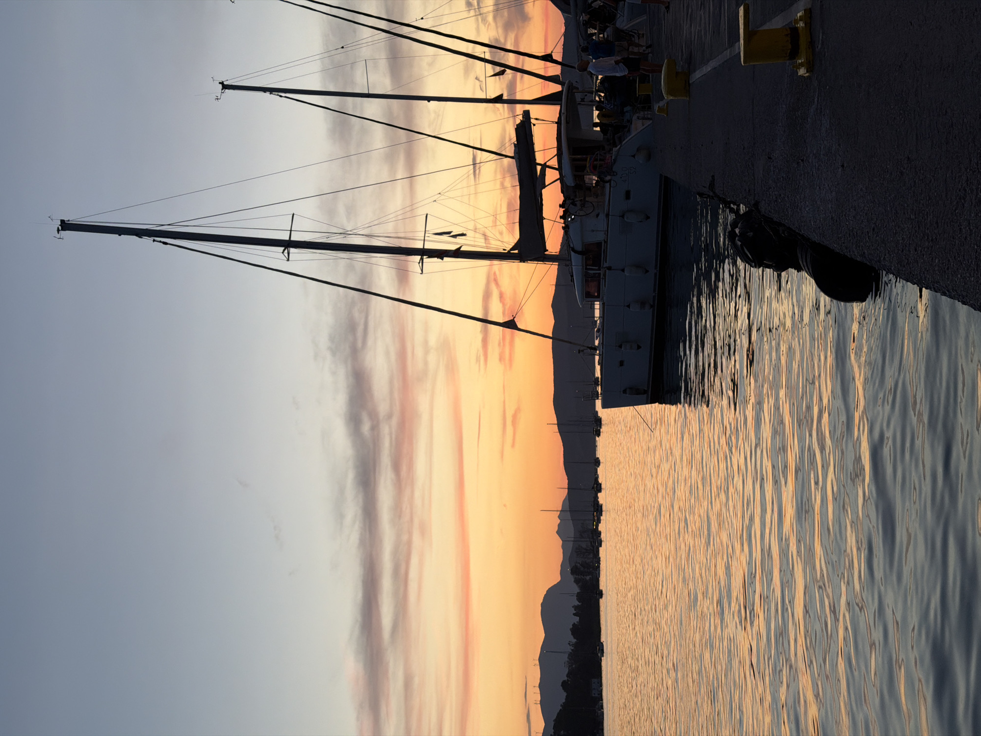 The image captures a serene marina scene at either sunrise or sunset, characterized by warm, vibrant colors. The sky, spanning from the upper left to the middle right, displays a beautiful gradient, transitioning from a soft light blue and grey at the top to rich oranges, pinks, and yellows near the horizon, with some streaky clouds catching the golden light.
In the foreground, the calm water of the marina reflects these vivid sky colors, creating shimmering patterns of light and shadow across its rippled surface. Along the right side of the frame, a dark-colored dock is visible, equipped with prominent yellow bollards. Several sailboats are moored to this dock, their tall masts and intricate rigging silhouetted against the brightly lit sky. The hull of a white or light-colored sailboat is prominent, reflecting some of the ambient light. On its side, the numbers "4.0 D 32" are faintly visible.
A few blurry figures, likely people, can be discerned on the dock next to the boats, suggesting activity or leisure. In the distance, across the horizon, a dark silhouette of mountains and land separates the glowing sky from the tranquil water. The overall atmosphere is peaceful and tranquil, highlighting the beauty of the twilight hour in what appears to be a coastal town or island setting, consistent with the location of Poros, Greece.