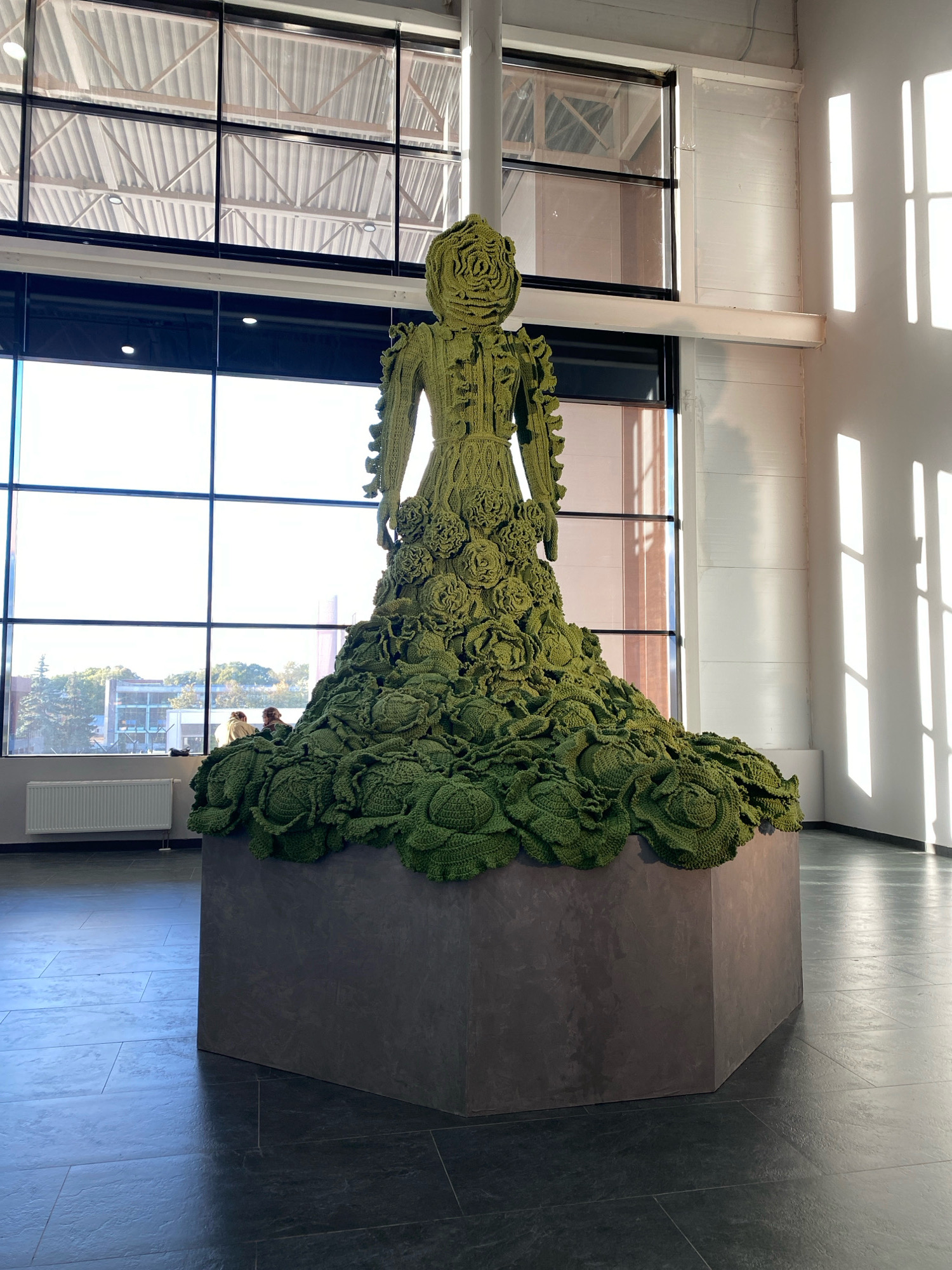 A large, green, crocheted sculpture of a woman is displayed in a bright, modern building with large windows. The sculpture is covered in crocheted rosettes and resembles a dress made of cabbage leaves. It stands on a dark, hexagonal pedestal. In the background, through the windows, trees and buildings are visible. Two people are seated near the sculpture, looking out the window. The floor is tiled with dark, large square tiles. The sunlight casts geometric shadows on the wall to the right. This artwork is on display at the Khodynka Gallery, located in Khimki, Russia.