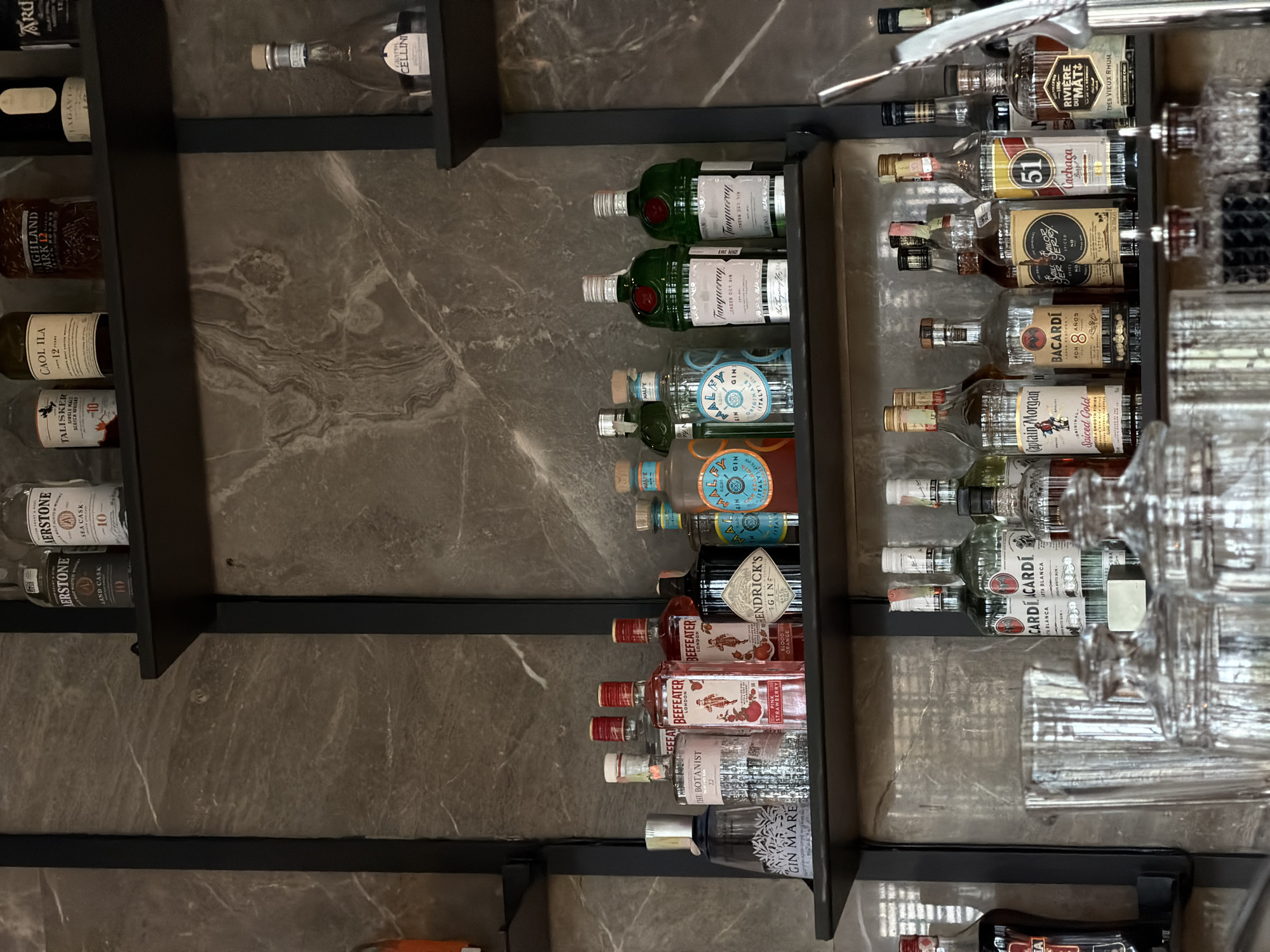 This image shows a bar with shelves stocked with various alcoholic beverages. Brands visible include gin such as Tanqueray, Hendrick's, Gin Mare, The Botanist, Beefeater; rum such as Bacardi, Captain Morgan; scotch whiskey such as Talisker, Caol Ila, Highland Park, and Aerstone. Also visible are cachaça 51, Riviere du Mat, and Cellini. There are many glasses on the right side of the image. The background walls and shelves are of dark gray and black color with marble patterns. The location is unknown and there is no indication in the image that the photo was taken in Bishkek City, Kyrgyzstan.