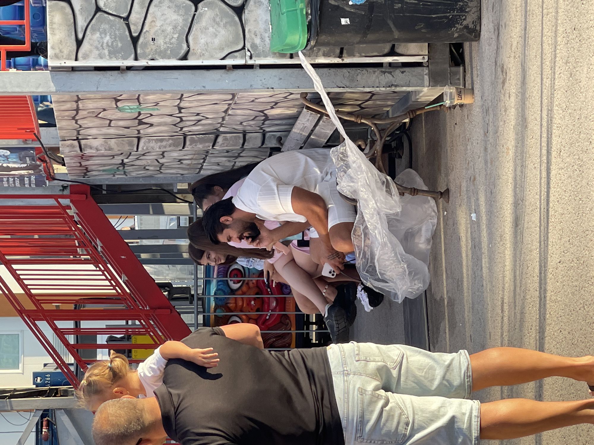 A group of people are gathered outdoors in what appears to be an amusement park or fairground, possibly in the city of Varna, Bulgaria. In the foreground, a man in a black t-shirt and light denim shorts stands with his back to the camera, carrying a young child on his shoulders. The child is wearing a white t-shirt. Behind him, a man in a white short-sleeved shirt and white shorts is sitting on a park bench, talking on his phone. He has a beard and tattoos on his arm. To his left, three young women are also seated on the bench. One woman in a pink shirt and another in a blue and pink top are looking in the same direction as the man on the phone. Another woman, partially visible, is wearing a pink top. The background features a red staircase, a wall decorated with a stone pattern, and colorful carnival imagery. A green trash bin and a black bin are also visible. A disposable coffee cup with a straw sits on the ground near the bench. The ground is paved with small stones, and shadows from structures create patterns on the surface.