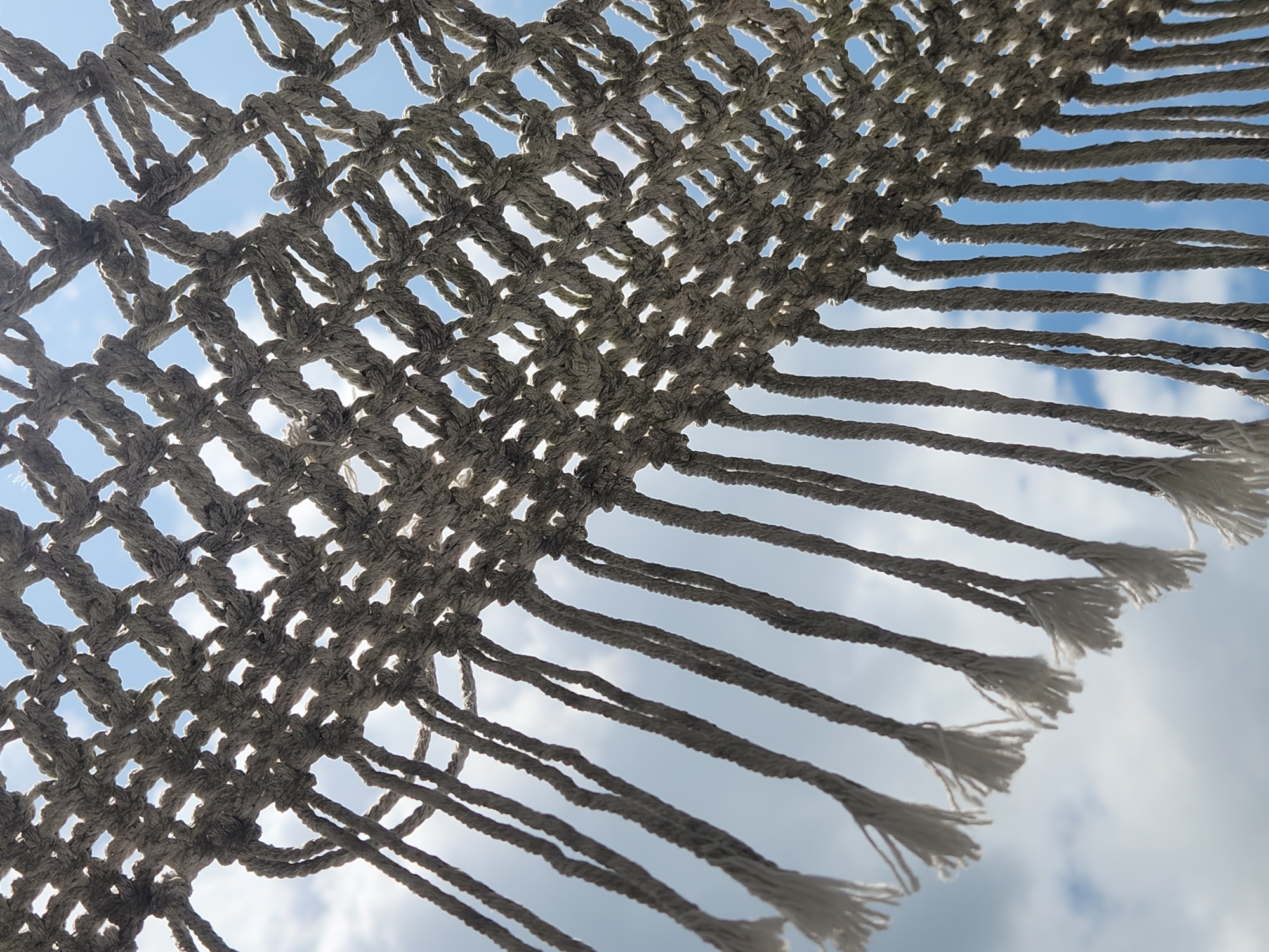 This is a low-angle, close-up photograph of a macrame net against a sky with clouds. The net is made of thick, woven rope and has a diamond pattern. The bottom edge of the net is fringed with hanging cords, some of which have tasseled ends. The sky behind the net is a pale blue with scattered white clouds, suggesting it is daytime. The sun is partially obscured by the net, casting shadows and highlighting the texture of the rope. There are no people or specific landmarks visible that would indicate a precise location within Maitland, United States of America. The scene appears to be outdoors, possibly a hammock or decorative net suspended in the air.