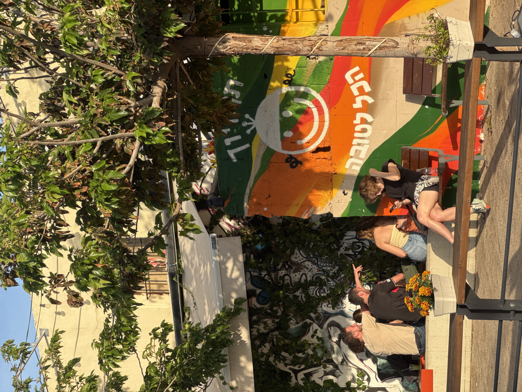 A sunlit outdoor scene in Tel-Aviv, Israel, presents a vibrant, graffitied wall with a colorful rainbow design and Hebrew text ("Hakol Letova" or "Everything is for the best"). In front of the wall, there are people seated or standing on a concrete bench. A woman in a black top and patterned shorts sits on the bench, possibly interacting with another person. Nearby, another person wearing a tan top and blue jean shorts stands. Adjacent to them, a couple stands together near a flower pot with orange blossoms. The scene is framed by a tree, and there are also a number of other plants in the area.