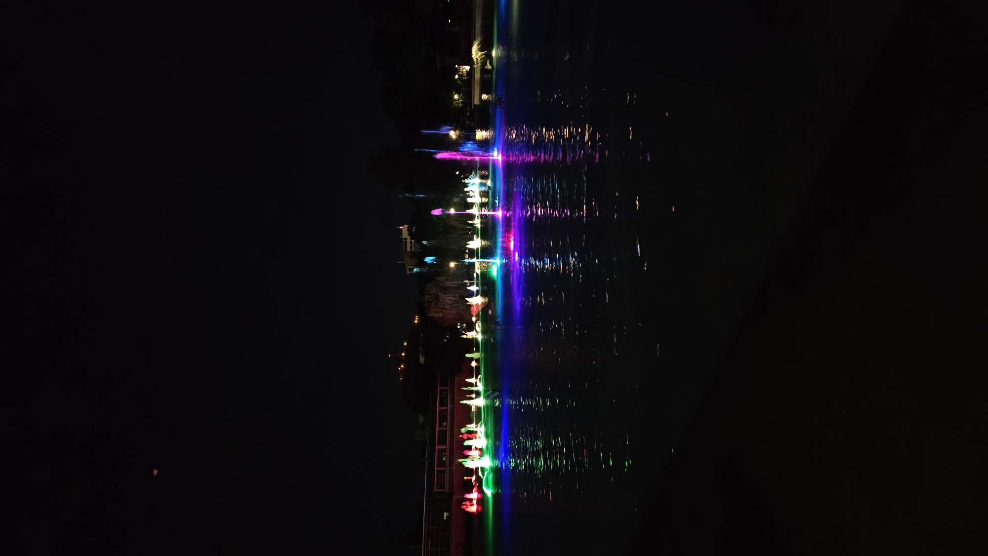 This image depicts a night view of a water fountain display, likely at a park or public space, with a dark sky overhead. The fountains are illuminated with vibrant, multi-colored lights, creating a visually striking effect as the water streams upwards and reflects on the dark water surface. In the background, there are some silhouetted trees and buildings, with lights indicating urban activity. The location is identified as the Tsar Simeon's Garden in Plovdiv, Bulgaria. The scene is serene and atmospheric, suggesting a public gathering or a place for evening leisure. There are no visible people in the immediate foreground, but the presence of chairs near some fountains suggests a seating area for viewers. The overall impression is one of a beautifully lit urban oasis at night.