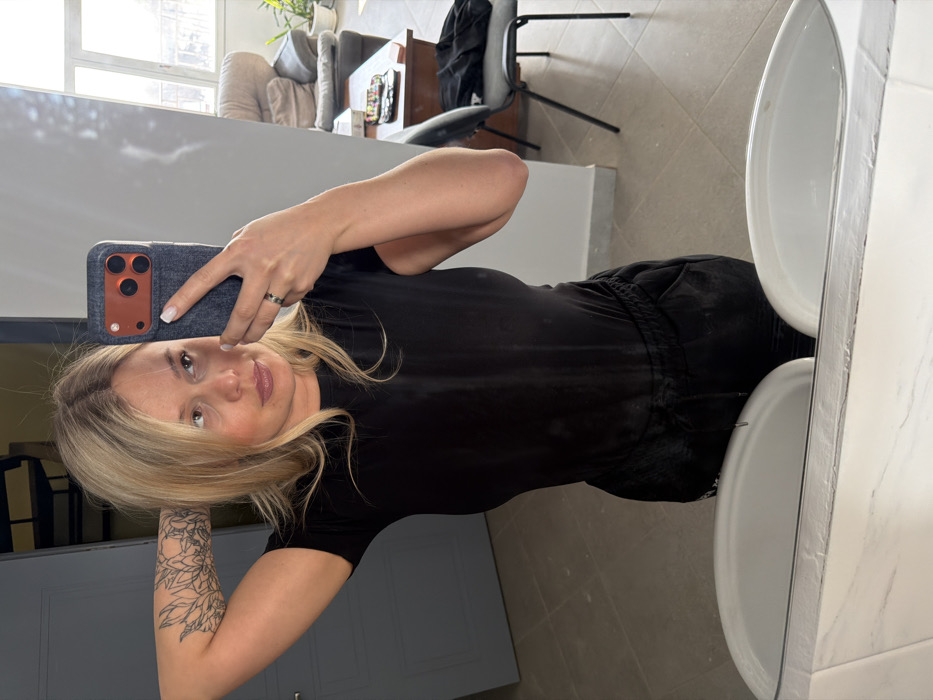 An individual with blonde hair is depicted taking a mirror selfie in an interior setting in Sarapul, Russia. The person, appearing female, wears a black short-sleeved top and dark, possibly black, drawstring pants. A black-ink floral tattoo is visible on her raised left upper arm. She holds a smartphone with a dark gray case and an orange camera module in her right hand, which has a ring on the ring finger.
The immediate foreground of the reflection shows two white oval sinks set into a white marbled countertop. The background on the left features a gray door and a dark, multi-level structure, possibly a staircase or shelving. The background on the right reveals a window admitting natural light, an upholstered armchair, a dark wooden table with various small objects, and a dark metal-framed chair with a black garment draped over it. A potted plant is also visible near the window. The floor is covered with light gray tiles.
