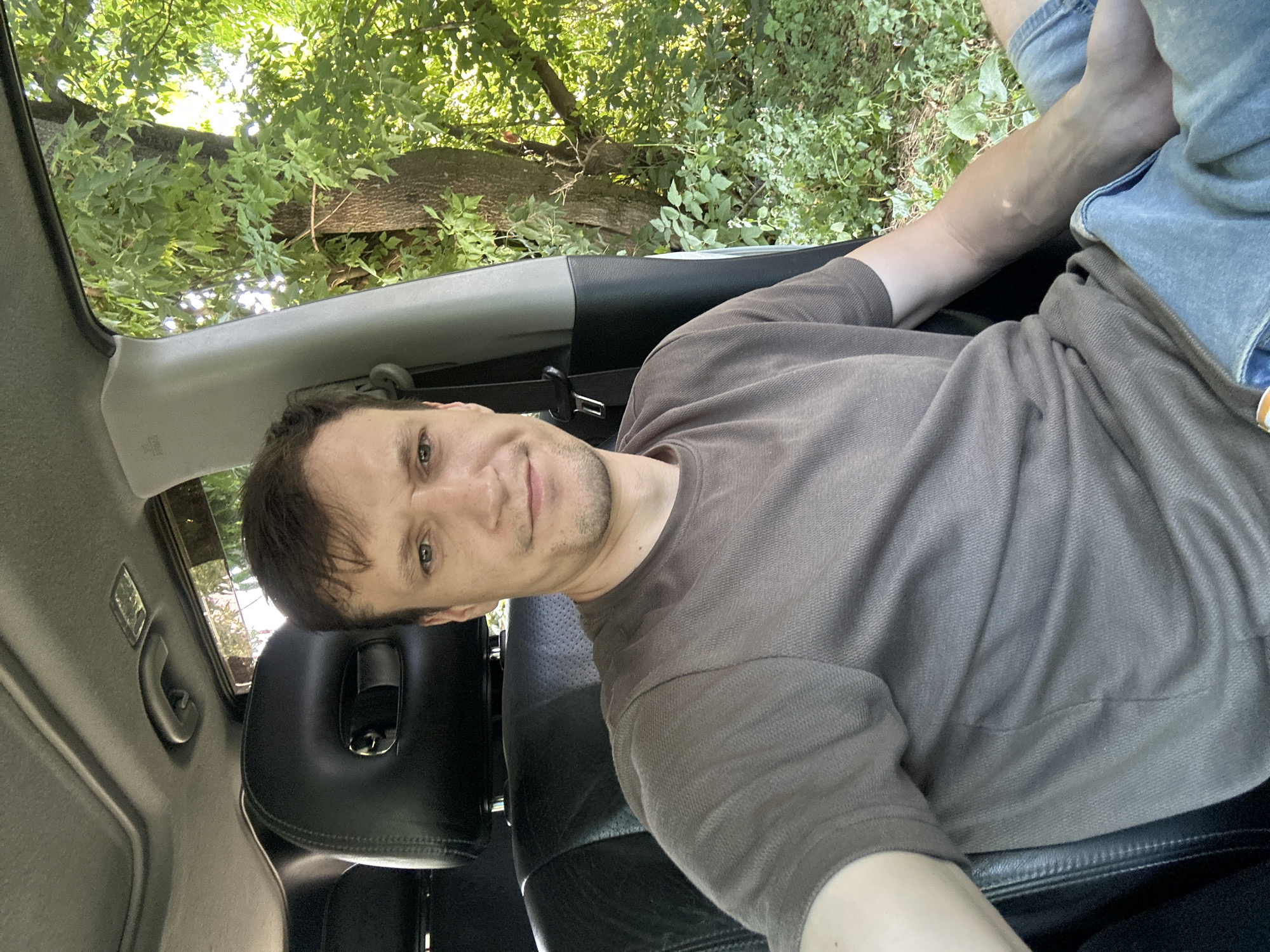 A man is sitting in the driver's seat of a car, with his right arm resting on the center console. The car is parked on a dirt road, with trees and foliage visible outside the open car doors. The man is wearing a brown t-shirt and blue jeans. The scene appears to be in a rural area, possibly on the outskirts of Makiv, Ukraine, given the dense vegetation and natural surroundings.