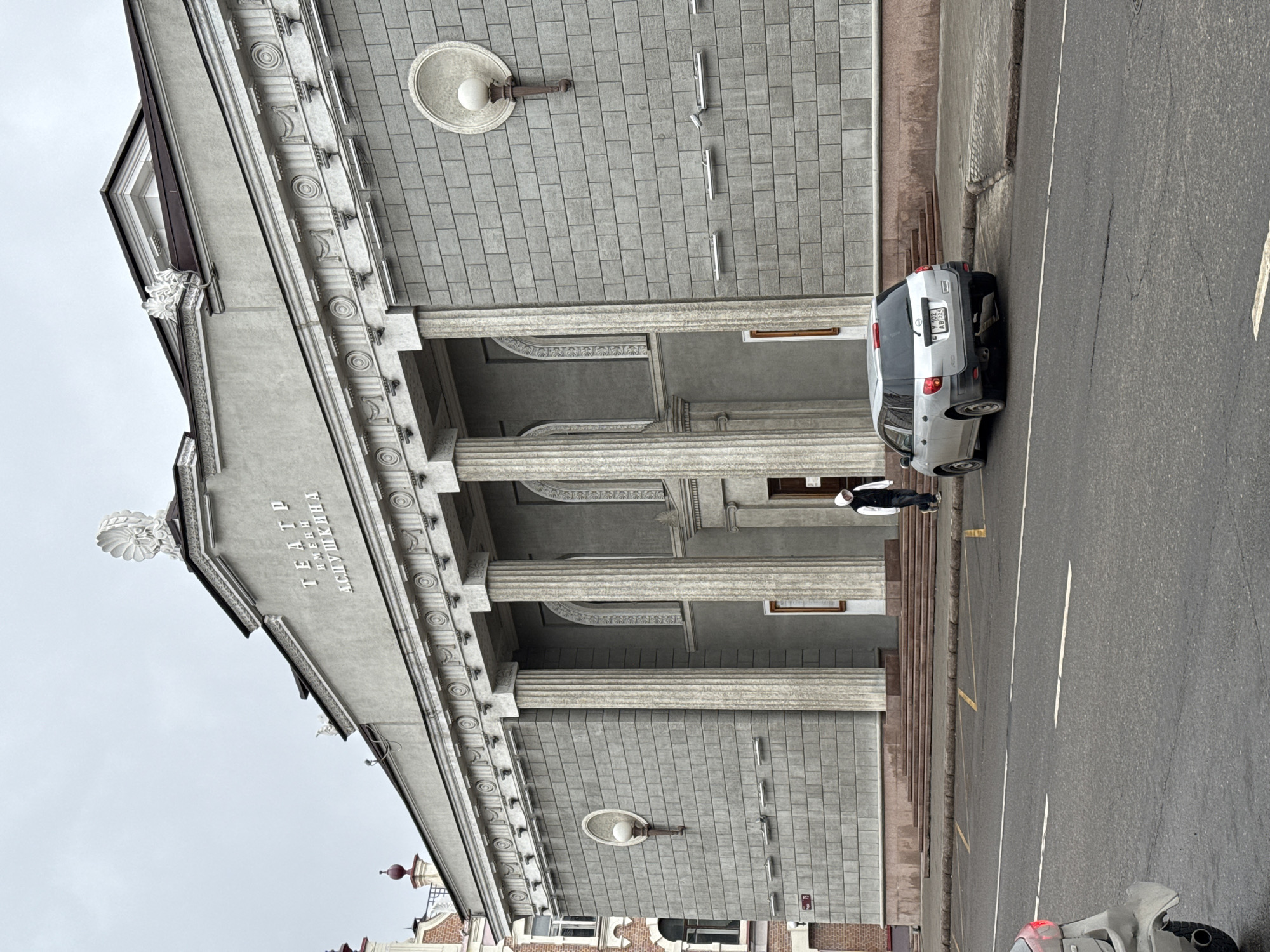 The image captures the facade of the Krasnoyarsk State Pushkin Drama Theater in Krasnoyarsk, Russia. The building is a gray, neoclassical structure with prominent columns and decorative elements. A silver car is parked on the street next to the theater's entrance stairs. A person in a dark vest and white hat is standing near the car. The street is paved with asphalt, and lane markings are visible. Other buildings are visible in the background, with some red brick elements. Overhead is a gray sky.
