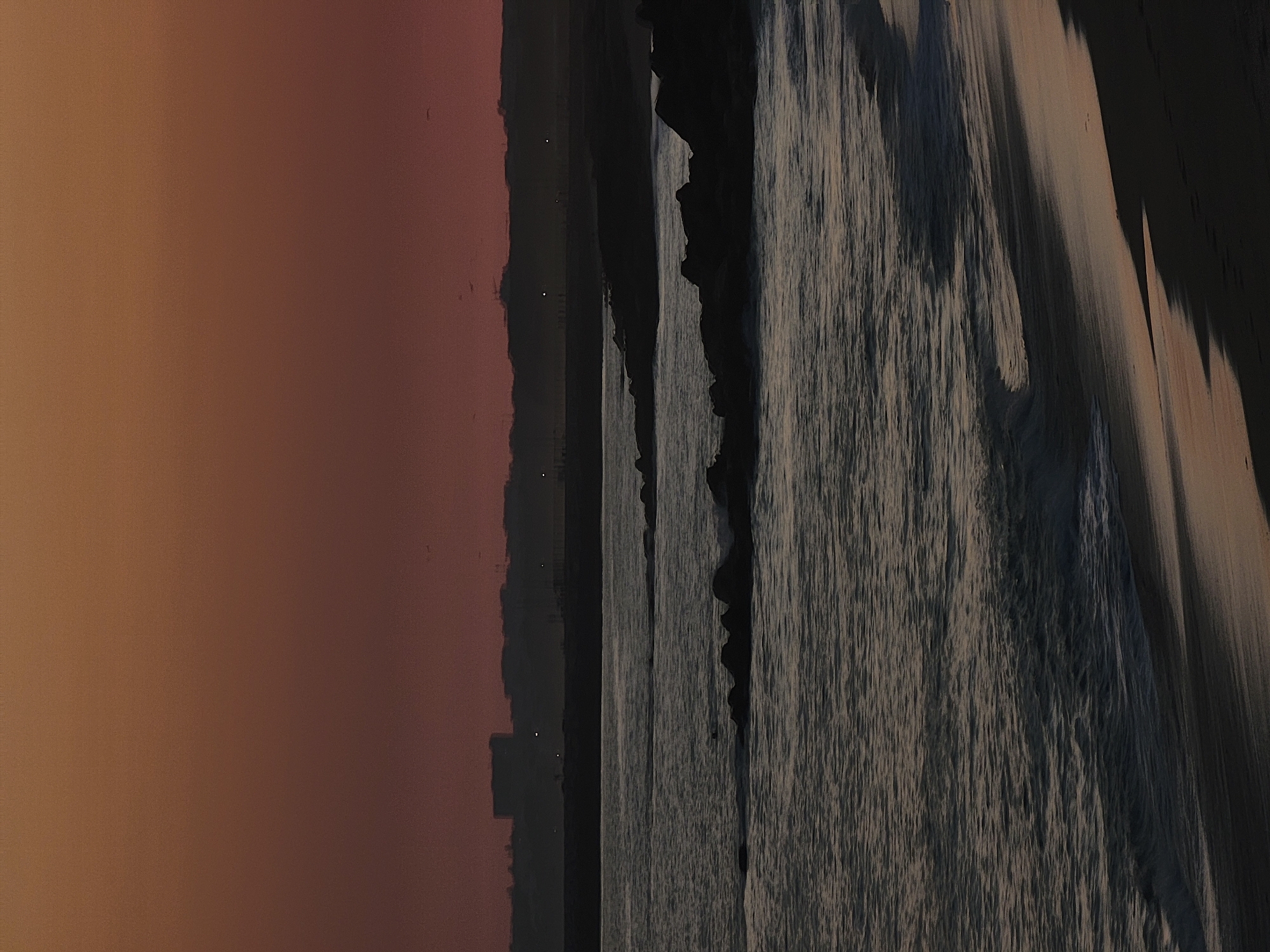 The image depicts a beach scene at dusk in Quarteira, Portugal. The composition is oriented vertically, with the left side dominated by a warm, gradient sky that transitions from a soft orange to a deep reddish-purple. This suggests either sunrise or sunset, but the rich colors lean towards sunset.
On the right side of the image, the ocean is visible, with waves creating a textured, almost striated pattern as they recede from the shore. The water appears calm but with a lively surface due to the movement of the waves. Dark, rocky formations jut out from the sea or are along the shoreline, creating a dramatic contrast with the water and the sky. Some of these formations appear jagged and steep.
In the distance, along the horizon line between the sky and the sea, there is a silhouette of buildings and structures, indicating an urban or developed area near the beach. A few faint lights are visible within this silhouette, adding to the sense of dusk. The overall mood is serene and contemplative, with the fading light casting long shadows and creating a peaceful atmosphere. There is no visible text in the image. The scene appears to be natural and tranquil, highlighting the beauty of the coastline as day turns into night.