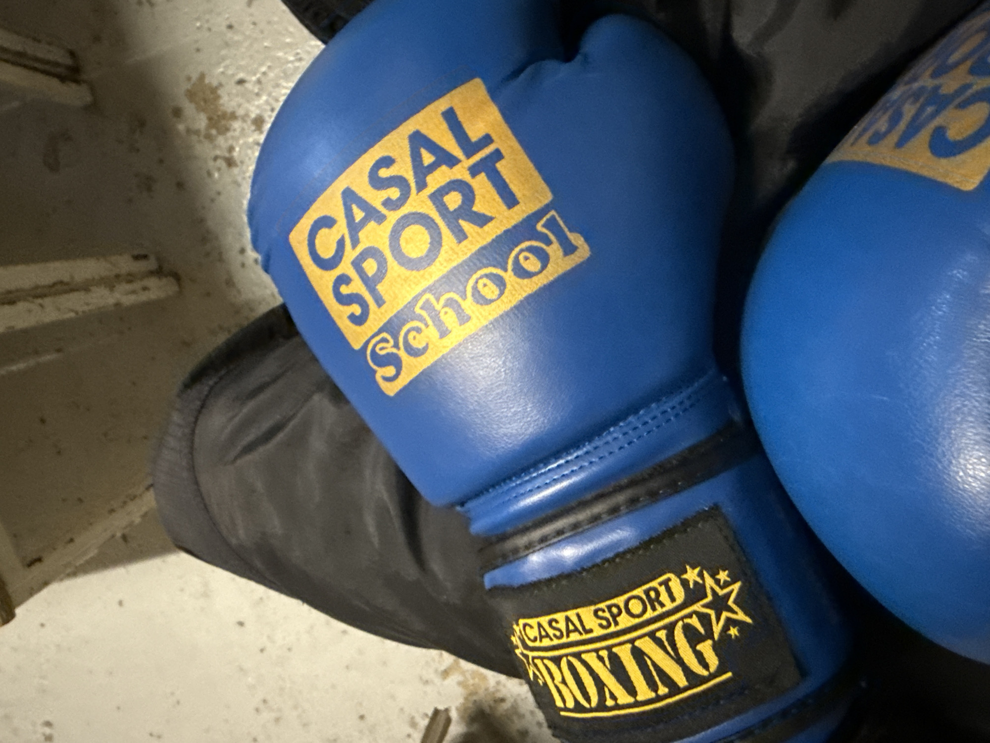 The image shows a pair of blue boxing gloves with yellow lettering that reads "CASAL SPORT School" on the upper portion and "CASAL SPORT BOXING" with stars on the wrist strap. The gloves appear to be resting on a dark-colored surface. The location is Sarcelles, France.