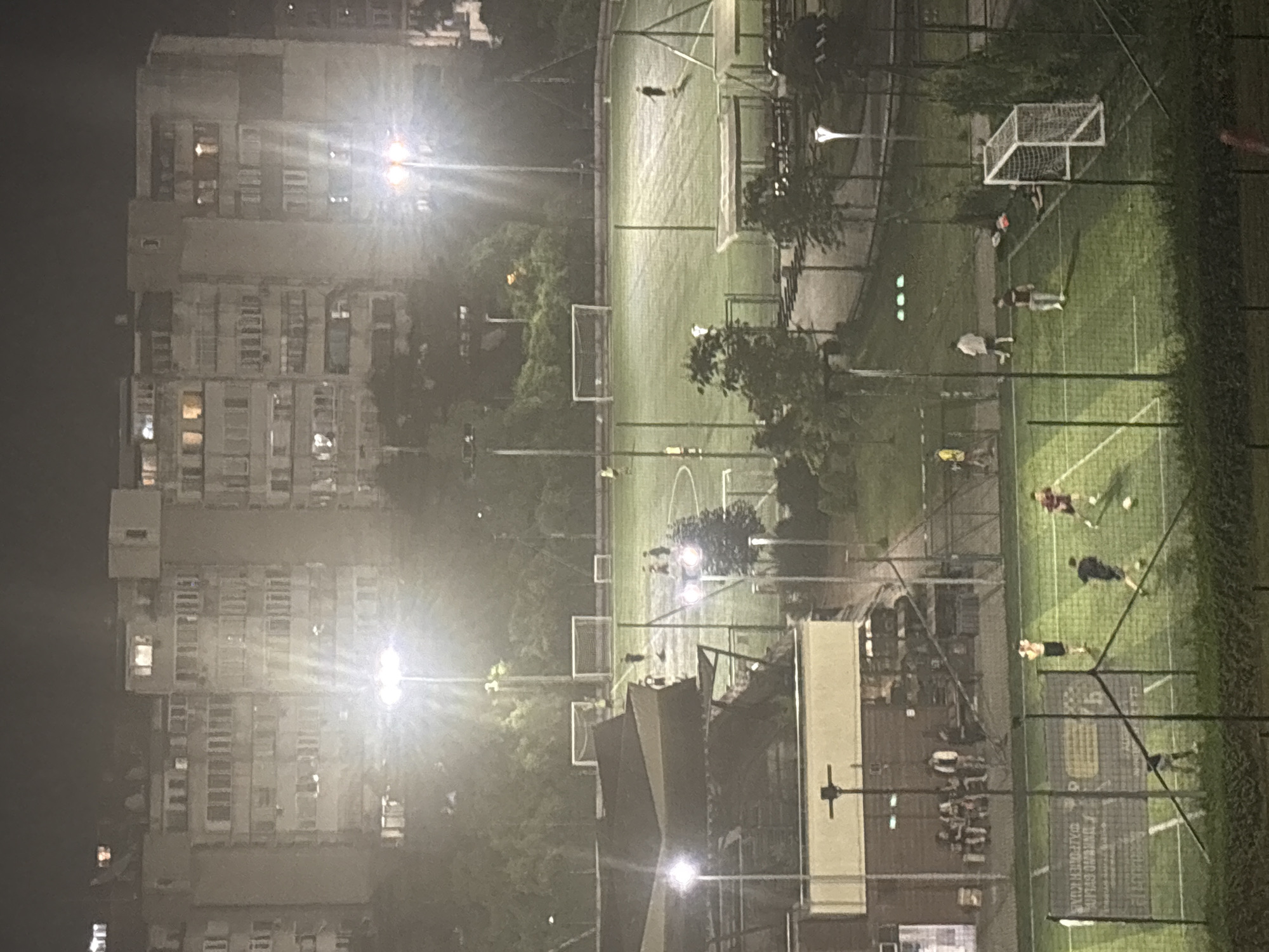 The image shows an aerial view of a brightly lit outdoor soccer field at night, with apartment buildings in the background. The soccer field is green and marked with white lines. Several people are playing soccer, some are standing or walking around the field. There are two soccer goals visible on the field. In the foreground, there is a fence and some trees. In the background, there are tall apartment buildings with lit windows. The overall scene depicts an urban recreational area at night.