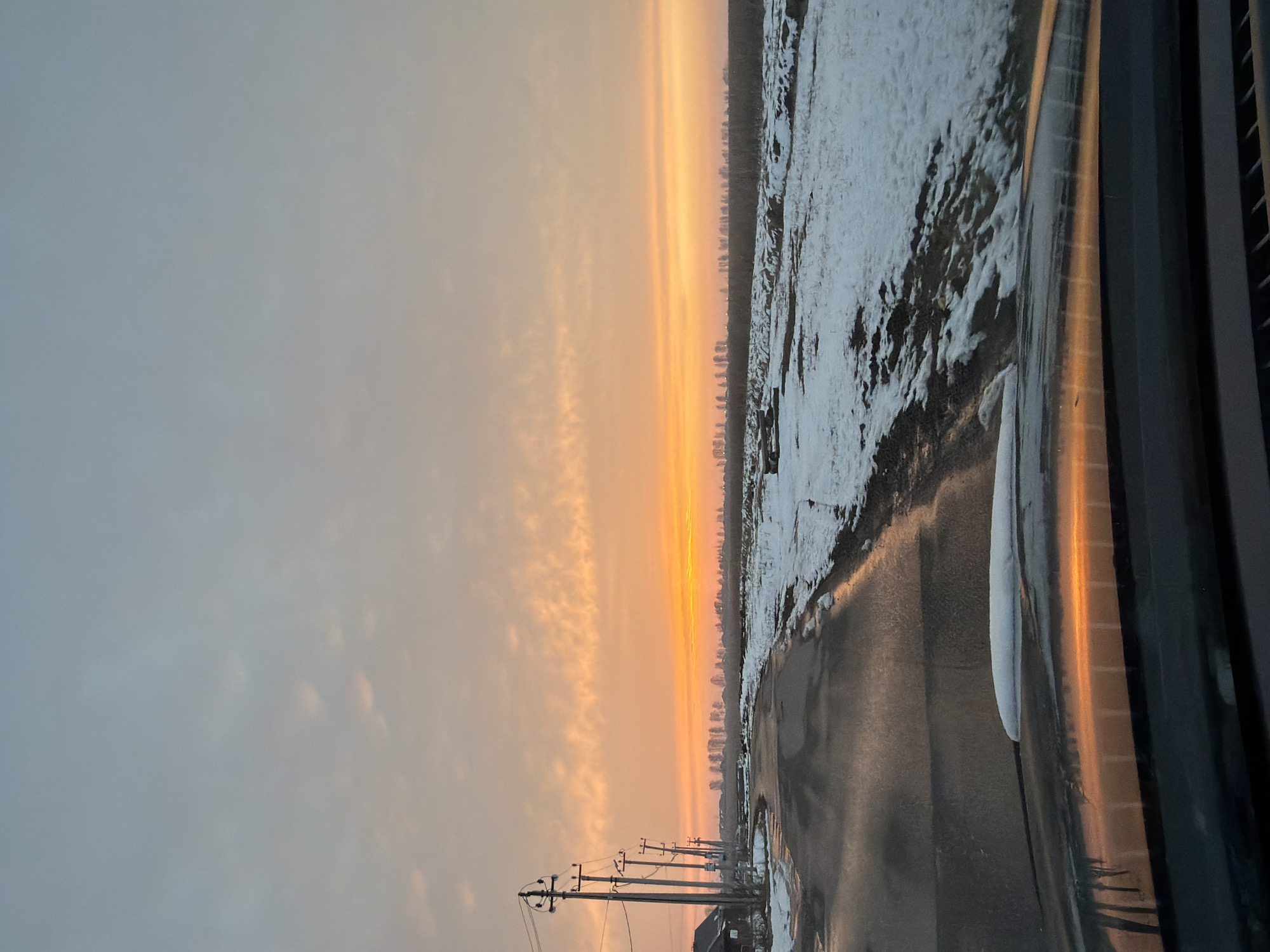 The image depicts a sunrise or sunset scene in what appears to be a rural or suburban area near Большая Шильна, Russia. A road runs through the lower part of the frame, reflecting light. Along the right side of the road, there is snow-covered terrain with vegetation visible in the background. In the distance, there are what appear to be buildings or structures suggesting a town or city. The sky is filled with orange and peach-colored light, indicating sunrise or sunset. Power lines are visible on the left side of the road. The photograph seems to be taken from inside a vehicle, with a portion of the vehicle's interior visible on the right side of the frame.