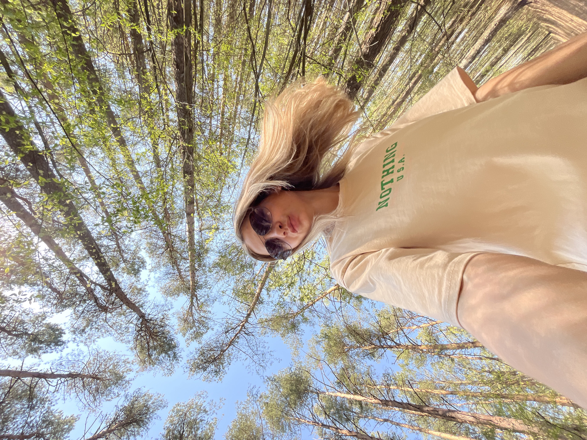 A low-angle, upward-facing shot in what appears to be a forest. The blue sky is partially visible through the canopy of trees. A woman with blonde hair and sunglasses is positioned in the foreground, wearing a beige t-shirt with the word "NOTHING" and "USA" printed on it. The location is Chaikovsky, Russia. The specific actions or interactions are limited as the image mainly focuses on the perspective and the woman's presence within the environment.