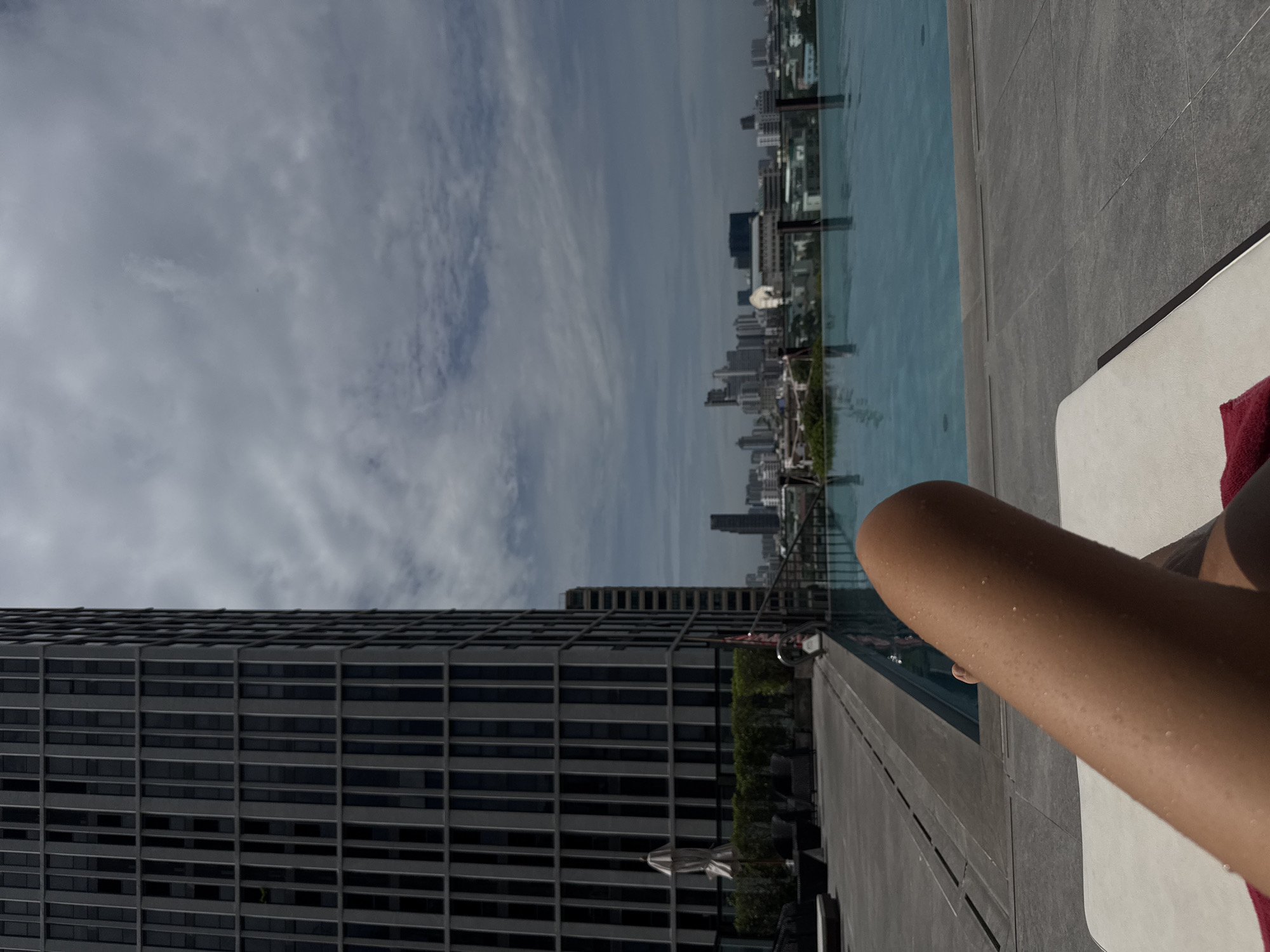 The image depicts a person's leg resting on a lounge chair near a rooftop infinity pool in Bangkok, Thailand. The leg appears wet, with water droplets visible on the skin, suggesting recent exposure to water. In the background, a modern high-rise building with a grid-like facade dominates the left side of the frame. Beyond the pool, the Bangkok skyline is visible, characterized by numerous buildings and a cloudy sky. The poolside area features grey paving stones, lounge furniture, and a hedge. The overall impression is one of relaxation and leisure in an urban setting.