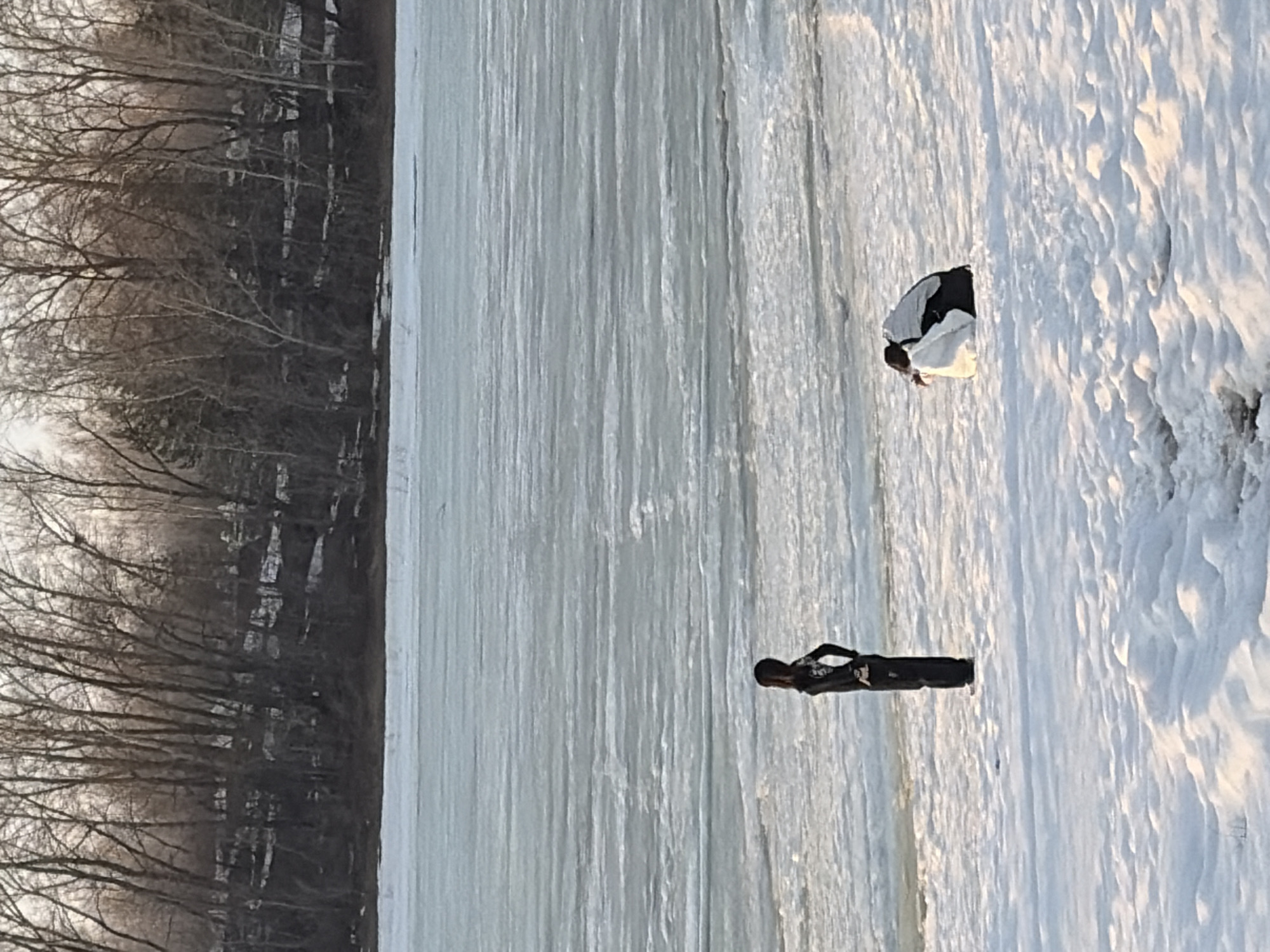 The image shows a winter scene in Chaikovsky, Russia. A frozen body of water, likely the Kama River, stretches across the frame, with snow-covered banks on one side. Bare trees are visible in the background. Two individuals are present. One person in dark clothing is standing upright near the edge of the ice, while another is bent over, possibly examining the ice or something on the surface. The sky is overcast, casting a diffuse light across the scene.