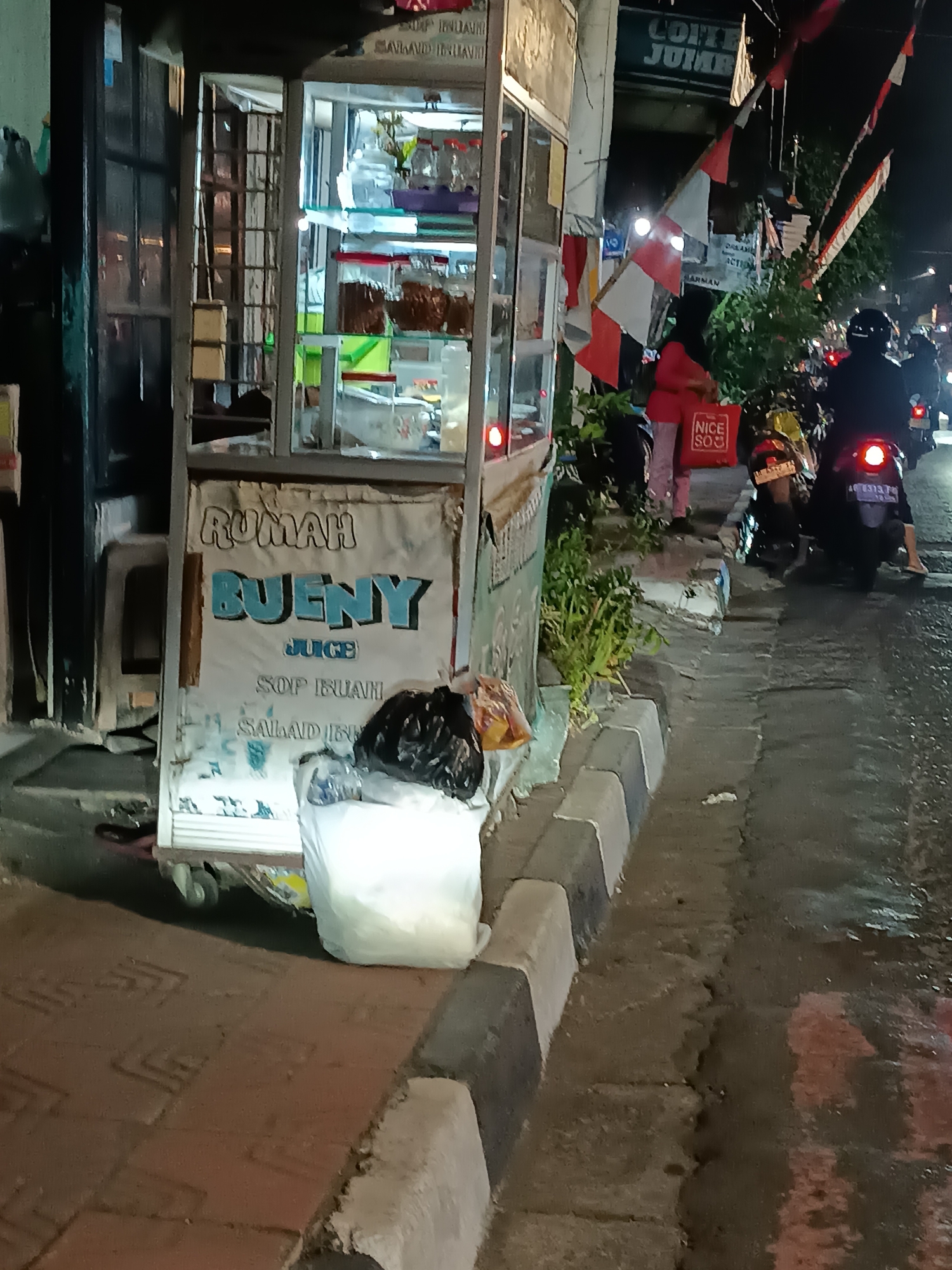 The image depicts a nighttime scene on a street in Yogyakarta, Indonesia, likely a bustling area with food vendors and traffic. In the foreground, a food cart is visible, featuring a sign that reads "RUMAH BUENY JUICE SOP BUAH SALAD BUAH". The cart appears to be a mobile unit, with wheels visible at the bottom. Two large white plastic bags are placed on the ground in front of the cart, with one partially containing a black trash bag.
Further back, a person in a pink shirt and dark pants stands near the cart, holding a red bag with the text "NICE SOJ". Next to them, two motorcycles are parked. The motorcycle on the right has a rider wearing a black helmet and jacket, and the motorcycle has a license plate. Behind the motorcycles, the street is visible with other vehicles, including a motorcycle with a rider also wearing a helmet. Red and white flags, possibly Indonesian flags, are displayed on poles in the background. The environment appears to be urban, with streetlights and building facades visible in the distance. The ground is paved with a combination of cobblestones and asphalt, and the road surface shows signs of being wet, possibly from recent rain.