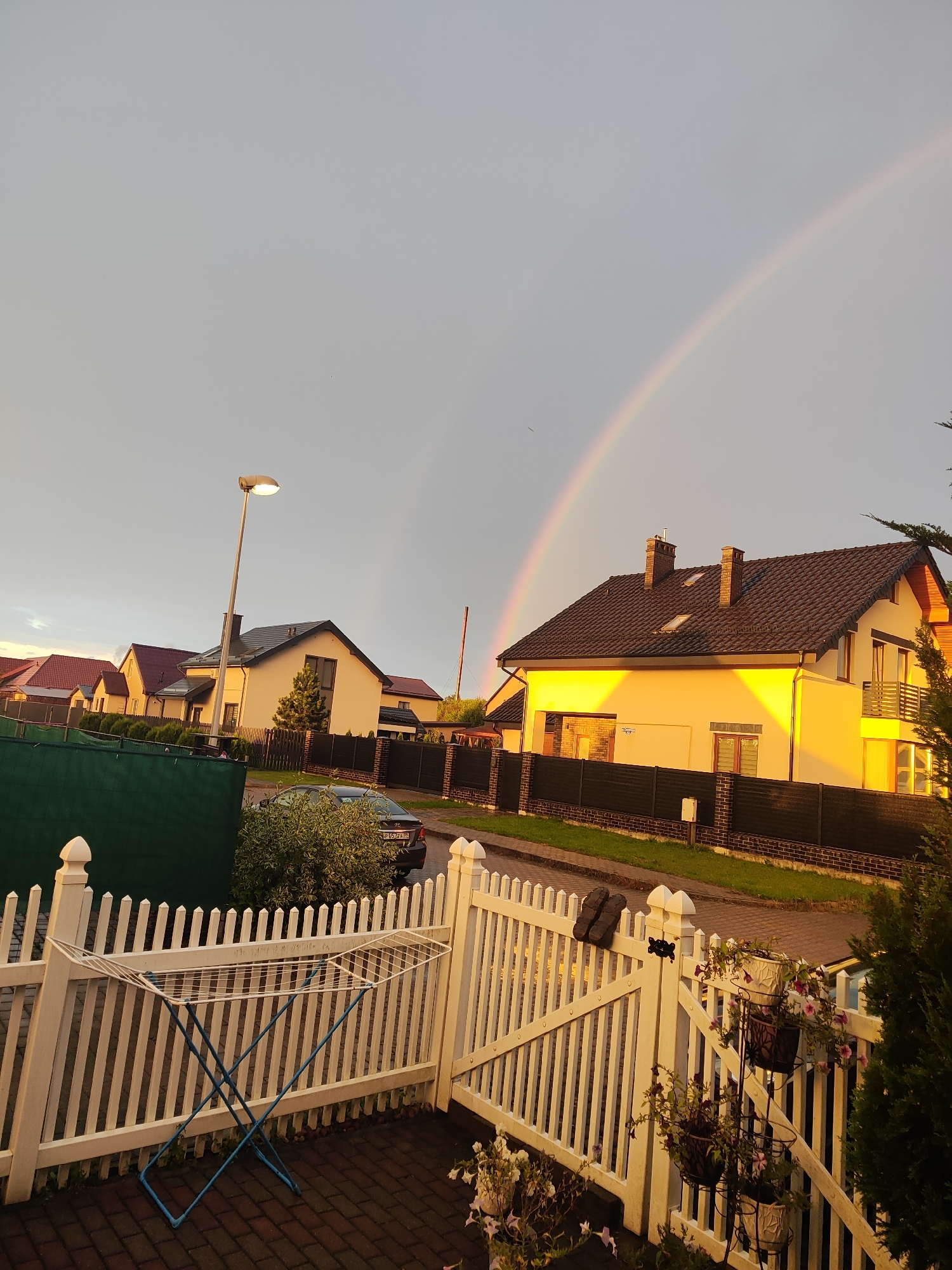 The image depicts a residential street scene in the city of Gvardeysk, Russia, at what appears to be either sunrise or sunset. A prominent double rainbow arcs across the overcast sky. In the foreground, a white picket fence encloses a paved area with a drying rack. A black sedan is parked on the street. Several houses with dark tiled roofs are visible, with one house in the middle ground illuminated by the warm light of the setting or rising sun, casting a golden glow on its facade. The environment is characteristic of a suburban neighborhood, with well-maintained homes and a street. No people are visible in the frame.
