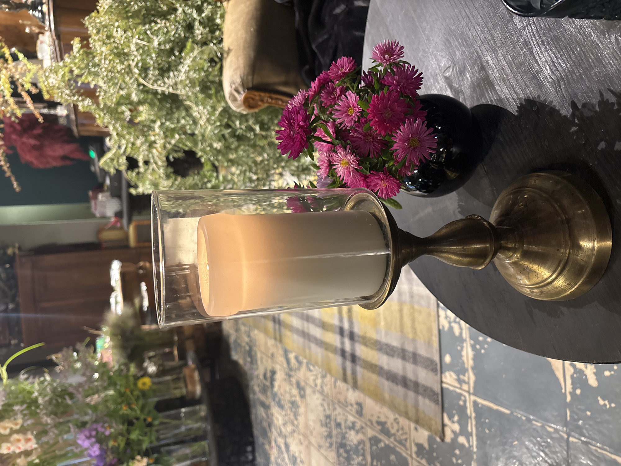 A lit candle in a glass hurricane sits on a brass candlestick on a dark, round table. To the right of the candlestick is a small black vase filled with a bouquet of pink asters. In the background, a checkered rug is visible on the floor, along with an antique cabinet and a plush armchair. The overall ambiance is cozy and inviting, suggesting a quiet moment in a dimly lit room. The scene is consistent with a setting in a cafe, restaurant, or a private residence in Riga, Latvia.