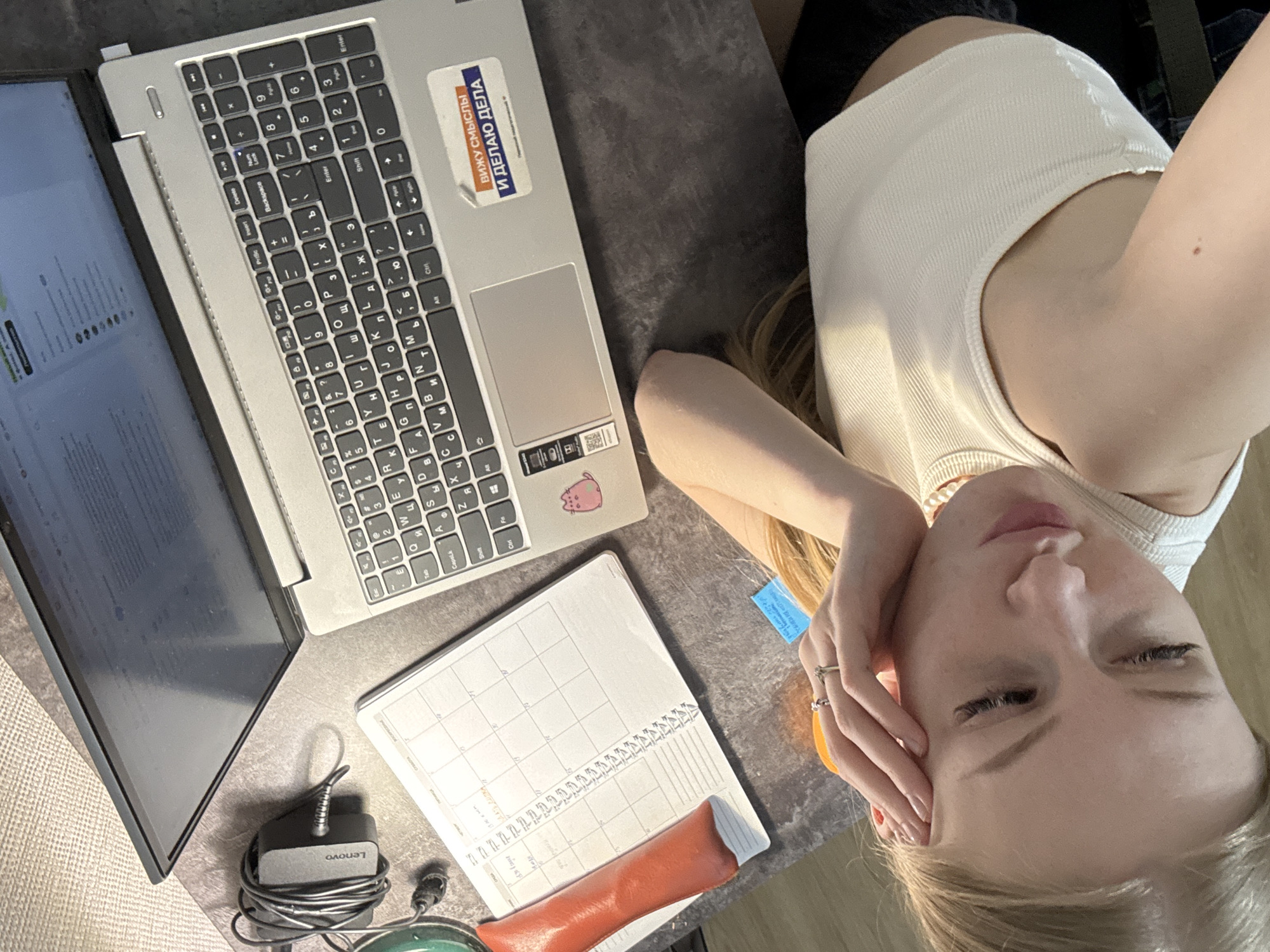 The image captures a top-down view of a young woman resting next to her workspace, likely in an indoor setting. The main subject is a woman with blonde hair, wearing a white ribbed tank top, lying on her side with her head supported by her left hand. Her eyes are partially or fully closed, conveying a sense of relaxation or contemplation.
In front of her, on a dark grey surface, is an open silver Lenovo laptop displaying text on its screen. The laptop's keyboard features both Latin and Cyrillic characters, indicating a Russian-speaking user. A notable detail on the laptop's palm rest is a small pink Pusheen cat sticker. Another sticker, placed above the keyboard, reads "ВИЖУ СМЫСЛЫ И ДЕЛАЮ ДЕЛА" (I SEE MEANINGS AND DO DEEDS), which further confirms the Russian context, consistent with the given location of Perm, Russia.
Next to the laptop is an open spiral-bound planner or calendar. On top of the planner rests an orange or reddish cylindrical object, possibly a pen case or a cosmetic item. A black Lenovo power adapter with a coiled cable is also visible near the laptop screen. A small blue sticky note is partially visible in the middle-right of the frame. The overall lighting suggests it is daytime, possibly with natural light illuminating the scene.