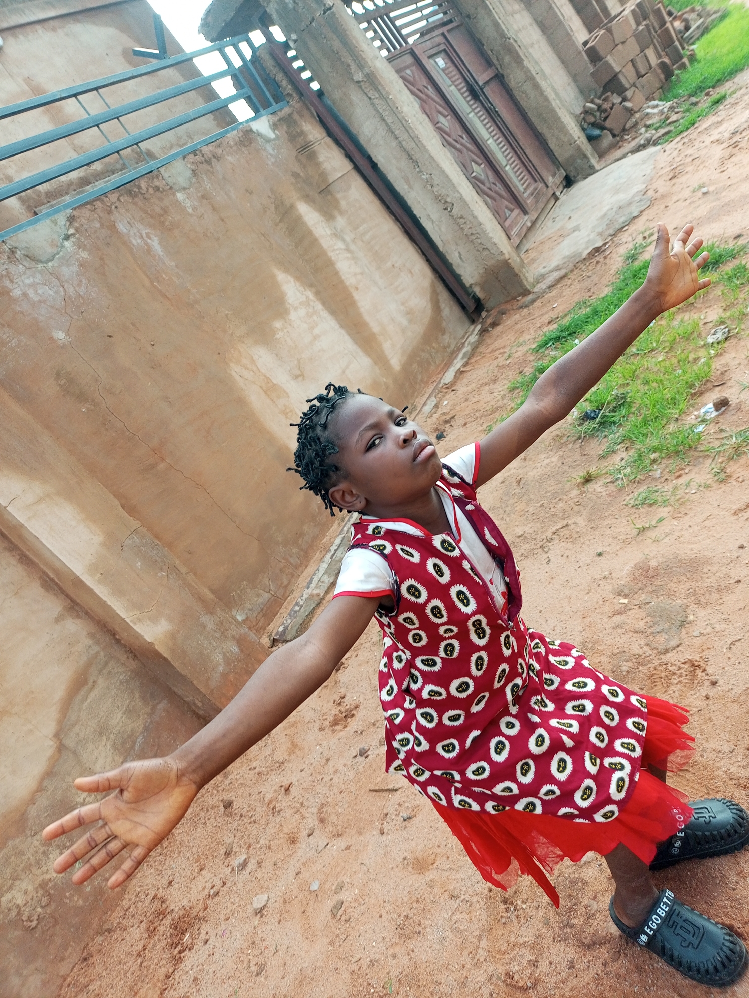 In Tempure, Nigeria, a young Black girl with braided hair is pictured outdoors. She is wearing a red and white patterned dress with a red underskirt, and black sandals. The girl is extending her arms outwards, with her left arm raised higher than her right. She is looking towards the camera with a serious expression. The background consists of a textured, light brown wall with some staining, a metal railing, and a brown metal gate. The ground is covered in reddish-brown dirt with some patches of green grass. The lighting suggests it is daytime.