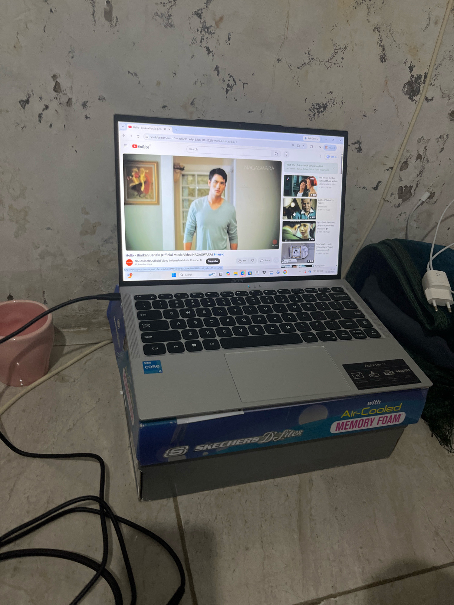 In Matesih, Indonesia, a laptop rests on a shoe box. The laptop is open and displays a YouTube video. The screen shows a man in a light blue shirt. The laptop's keyboard is visible, and a power cord is plugged into it. A pink mug is to the left of the laptop, and a white power adapter is on the right. The background is a textured wall.