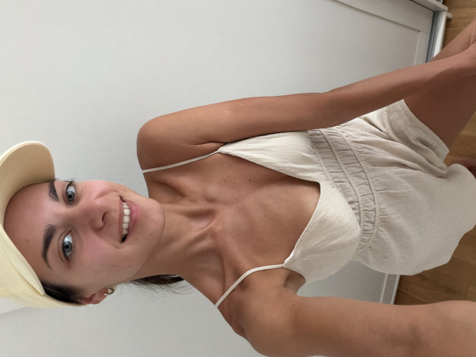 A woman wearing a beige romper and a light yellow baseball cap poses for a selfie against a plain white wall. She has fair skin, green eyes, and dark hair pulled back into a ponytail. She is smiling and looking at the camera. Her left arm is extended, holding the phone. The background is simple, with a wooden floor visible at the bottom and a white door on the right side. No specific landmarks of Budva, Montenegro are visible in the image.
