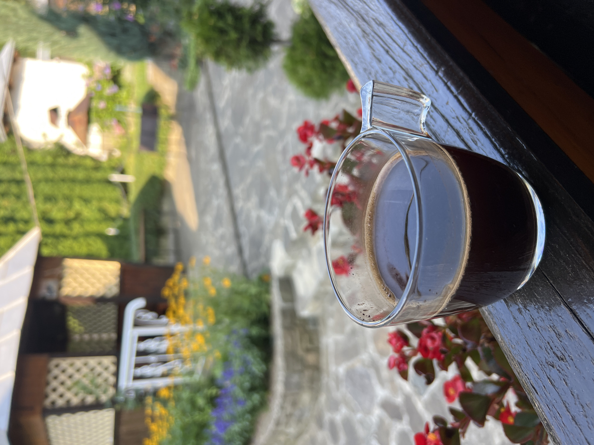 A close-up shot features a clear glass mug of dark coffee positioned on a weathered wooden railing. In the background, a charming, blurred courtyard unfolds. To the left, a rustic wooden gazebo with decorative latticework stands beside a raised garden bed filled with vibrant yellow and purple flowers. A white railing separates the gazebo from the flowers. Further into the background, a stone pathway leads through a sun-drenched garden. Greenery and hints of other structures are visible, suggesting a tranquil outdoor setting. The overall scene evokes a peaceful morning in Yaremche, Ukraine, with the coffee serving as the focal point against the soft-focus backdrop of the garden.