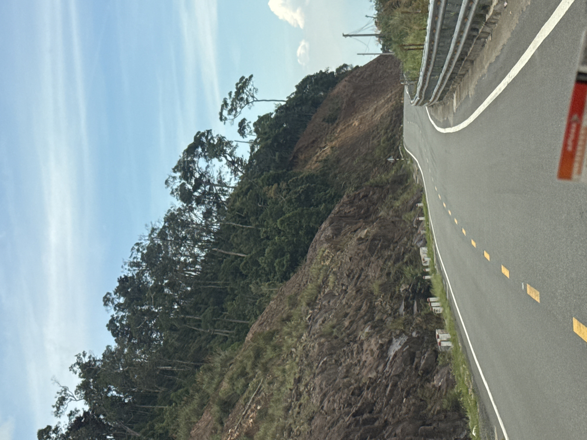 The image depicts a winding asphalt road passing through a mountainous area with steep, rocky slopes and dense vegetation. The road is marked with yellow dashed lines and a white solid line on the right side. Guardrails are present along the right edge of the road. Tall trees with sparse foliage grow along the top of the slopes, silhouetted against a light blue sky with scattered white clouds. In the foreground, a portion of a red object with white text is visible, partially obscuring the view of the road. The scene appears to be a remote, scenic route, likely in a rural area. The location is identified as Nam Khanh Vinh, Vietnam.